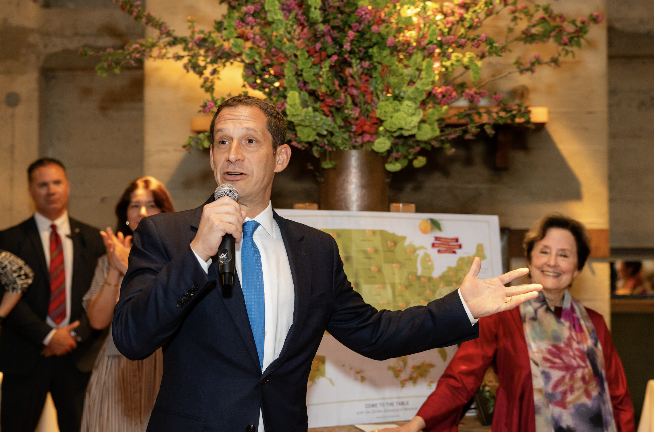 A man in a dark suit with a blue tie holding a microphone and speaking to an audience during a gathering. Several people, including a woman in red and others, are standing around him. A large floral arrangement with pink, purple, and green flowers an