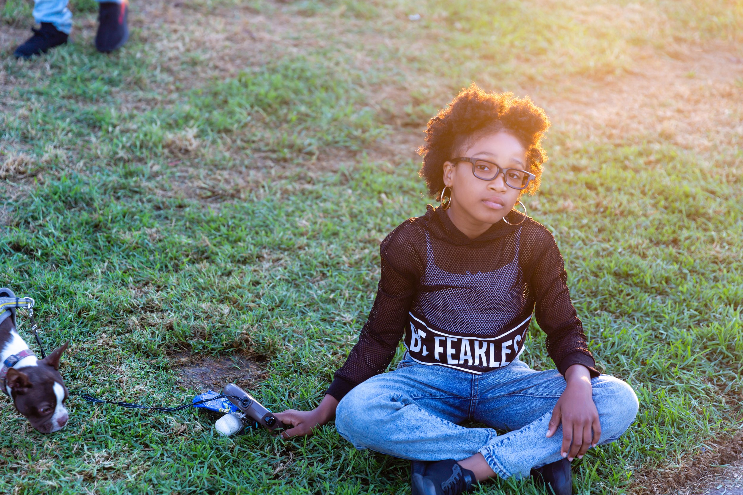 A young woman with glasses and hoop earrings sitting on grass outdoors during golden hour, wearing a black mesh top with the words 'BE FEARLESS'. A small dog with a brown and white coat is on a leash near her.