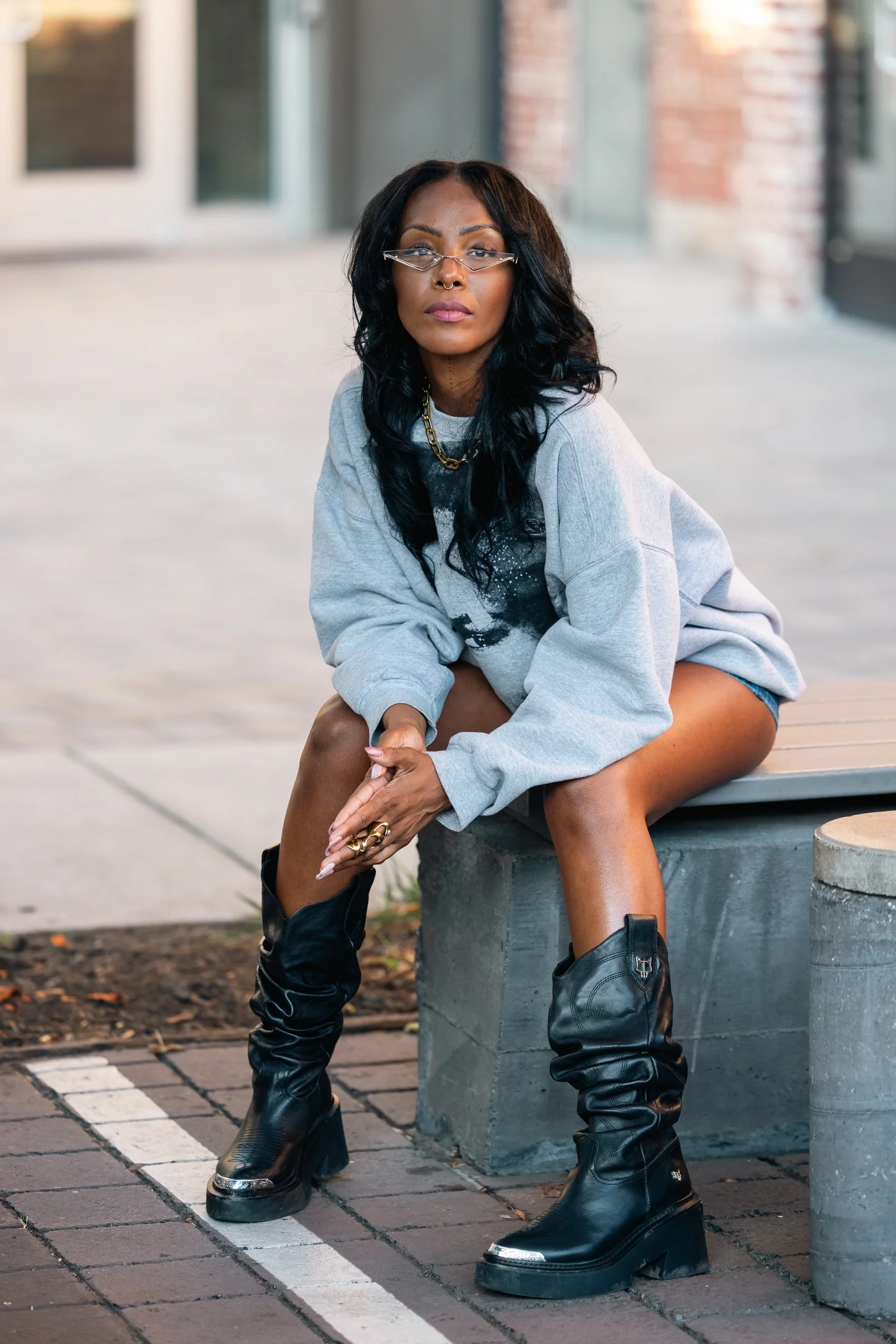 A woman with long black wavy hair, wearing glasses, a grey oversized sweatshirt, shorts, and black platform boots, sitting on a concrete bench outdoors.