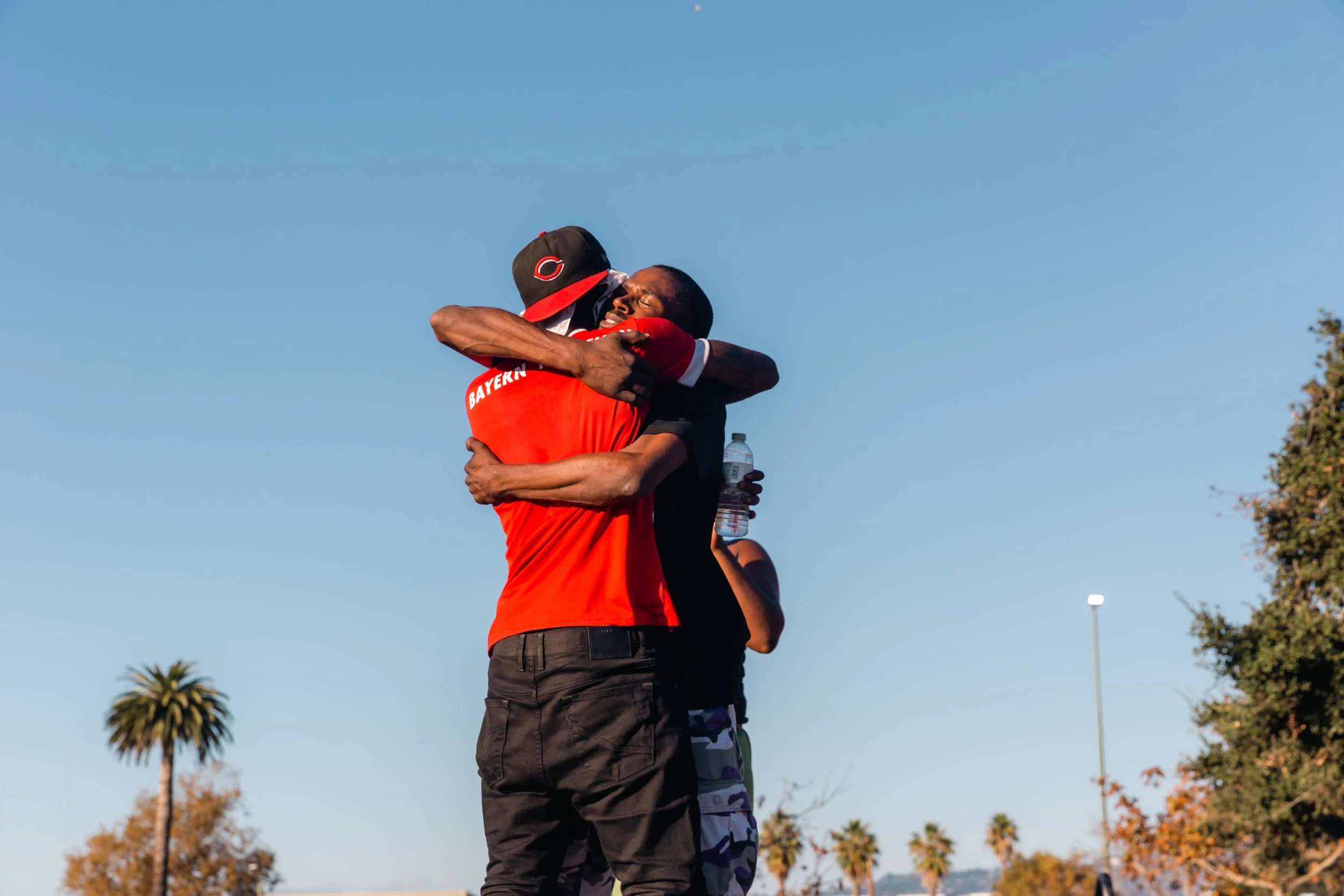 Two men hugging outdoors under a clear blue sky, with palm trees and other trees in the background. One man is wearing a baseball cap with a Cincinnati Reds logo and a red shirt, while the other is in a black shirt. One is holding a water bottle.