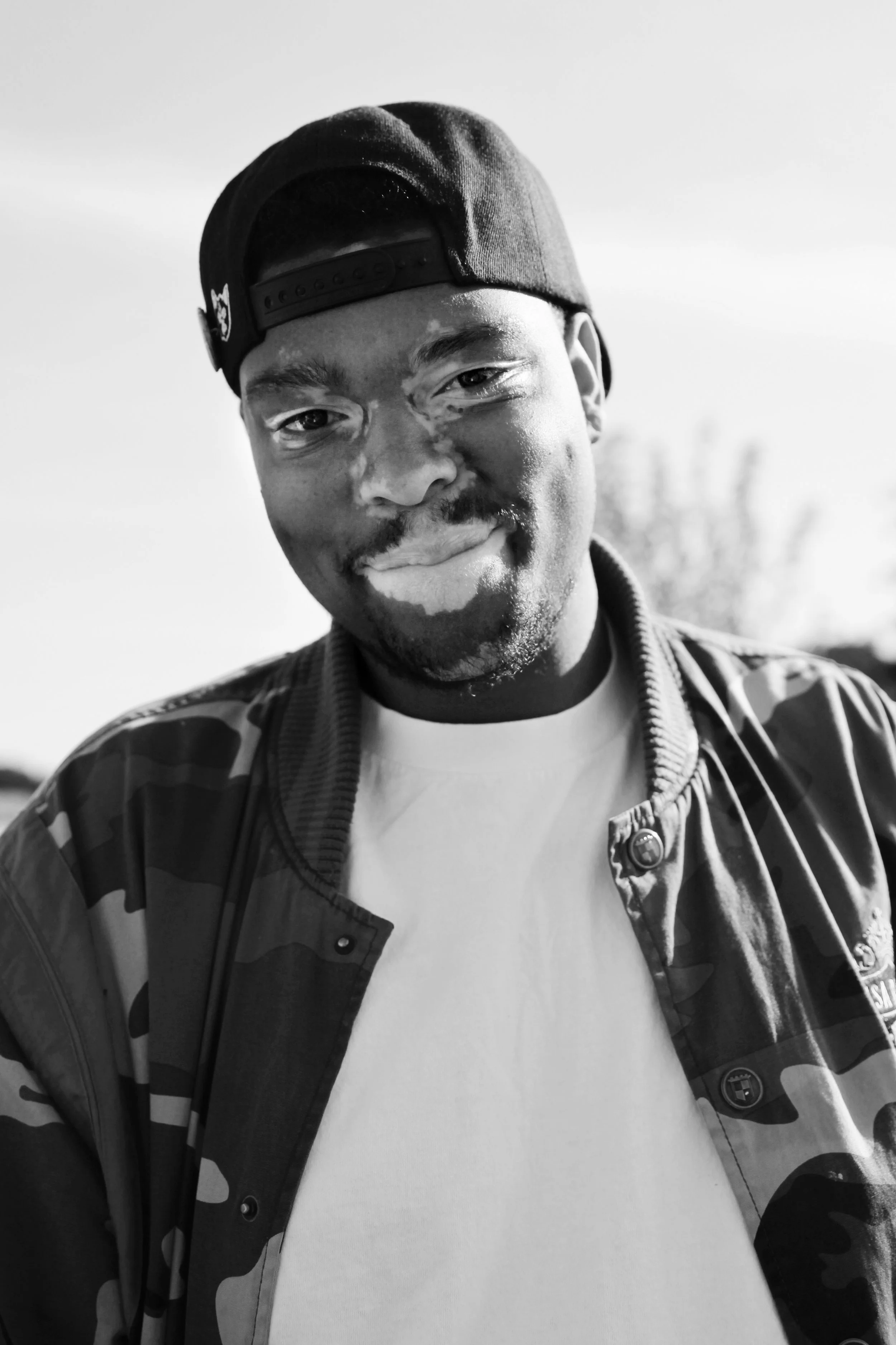 A young man with a mustache and beard wearing a black baseball cap backwards, a camouflage jacket, and a white T-shirt, smiling outdoors.