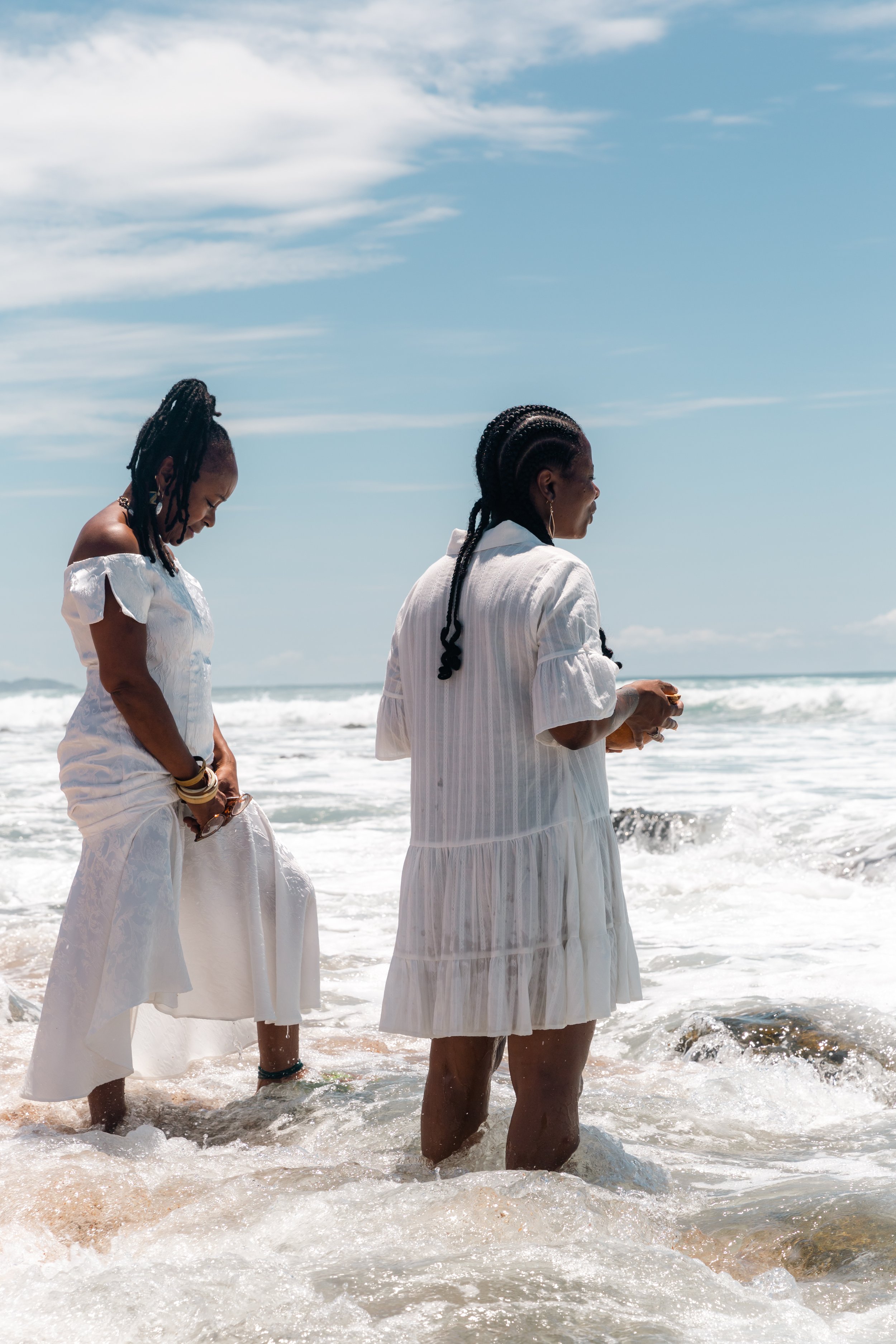 Three women wearing white dresses standing in the shallow water of the beach with waves, under a partly cloudy sky.
