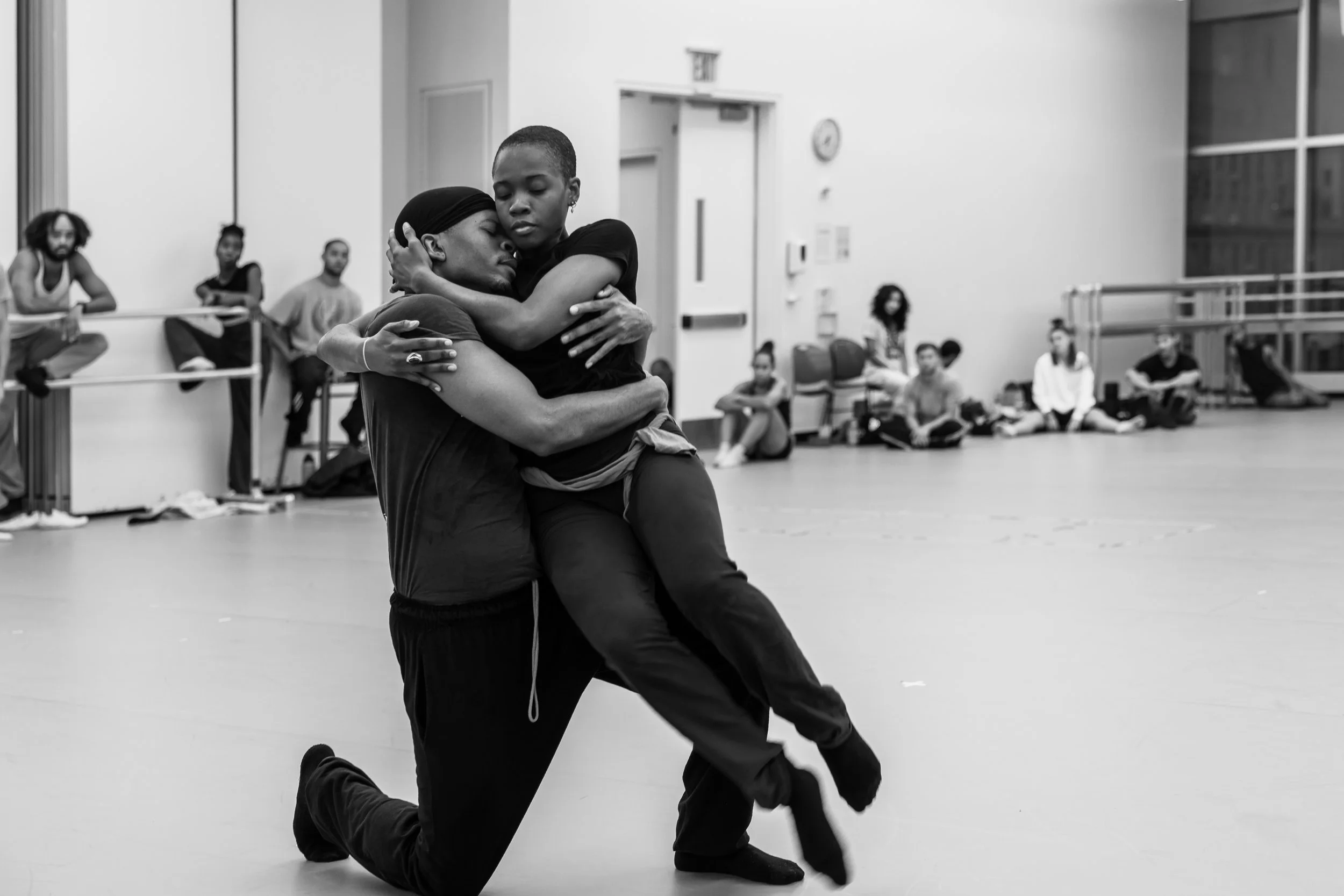 Two people dancing closely in a dance studio, surrounded by other dancers sitting and watching in the background.