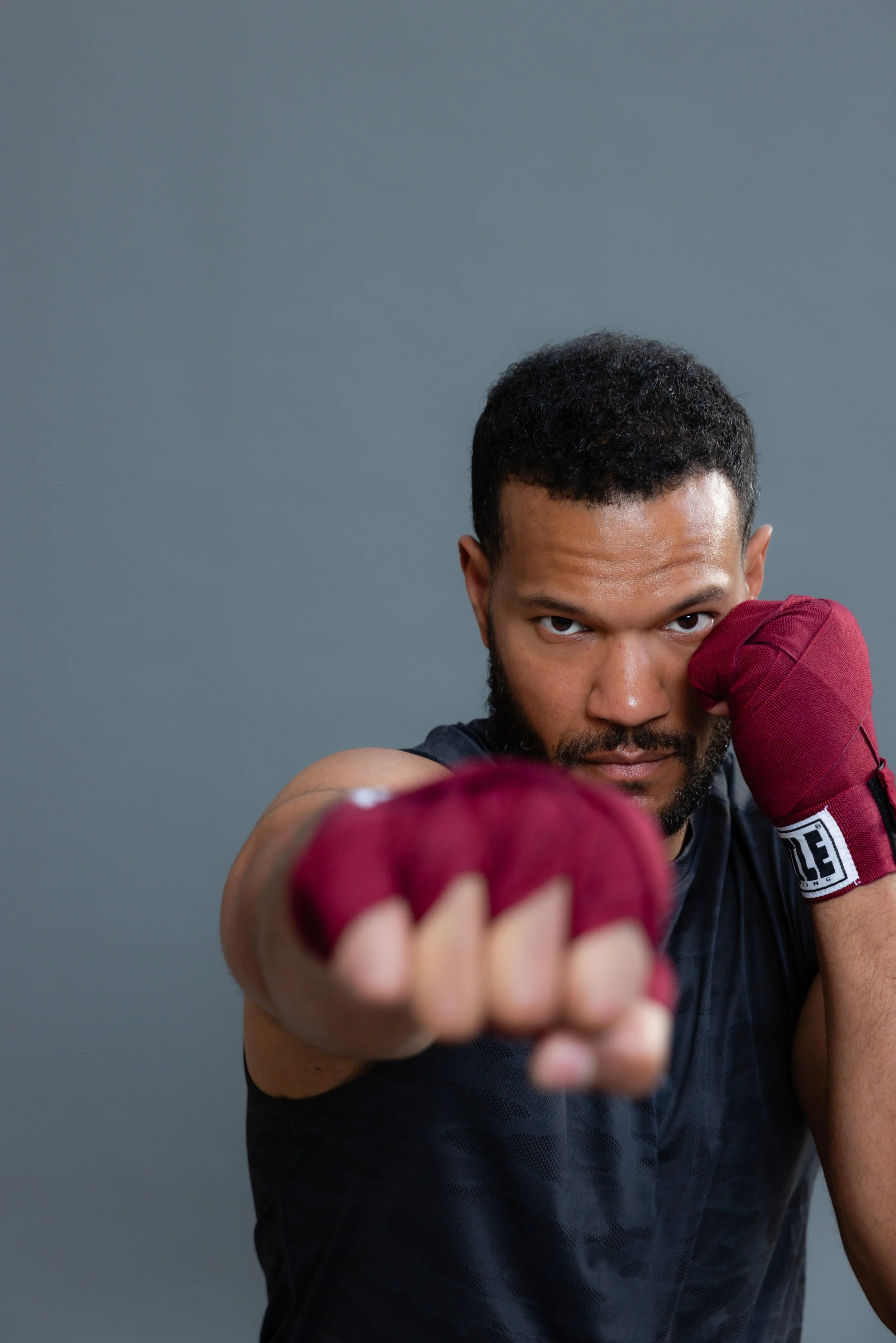 Man in a black shirt with reddish boxing gloves in a fighting stance, looking directly at the camera against a gray background.