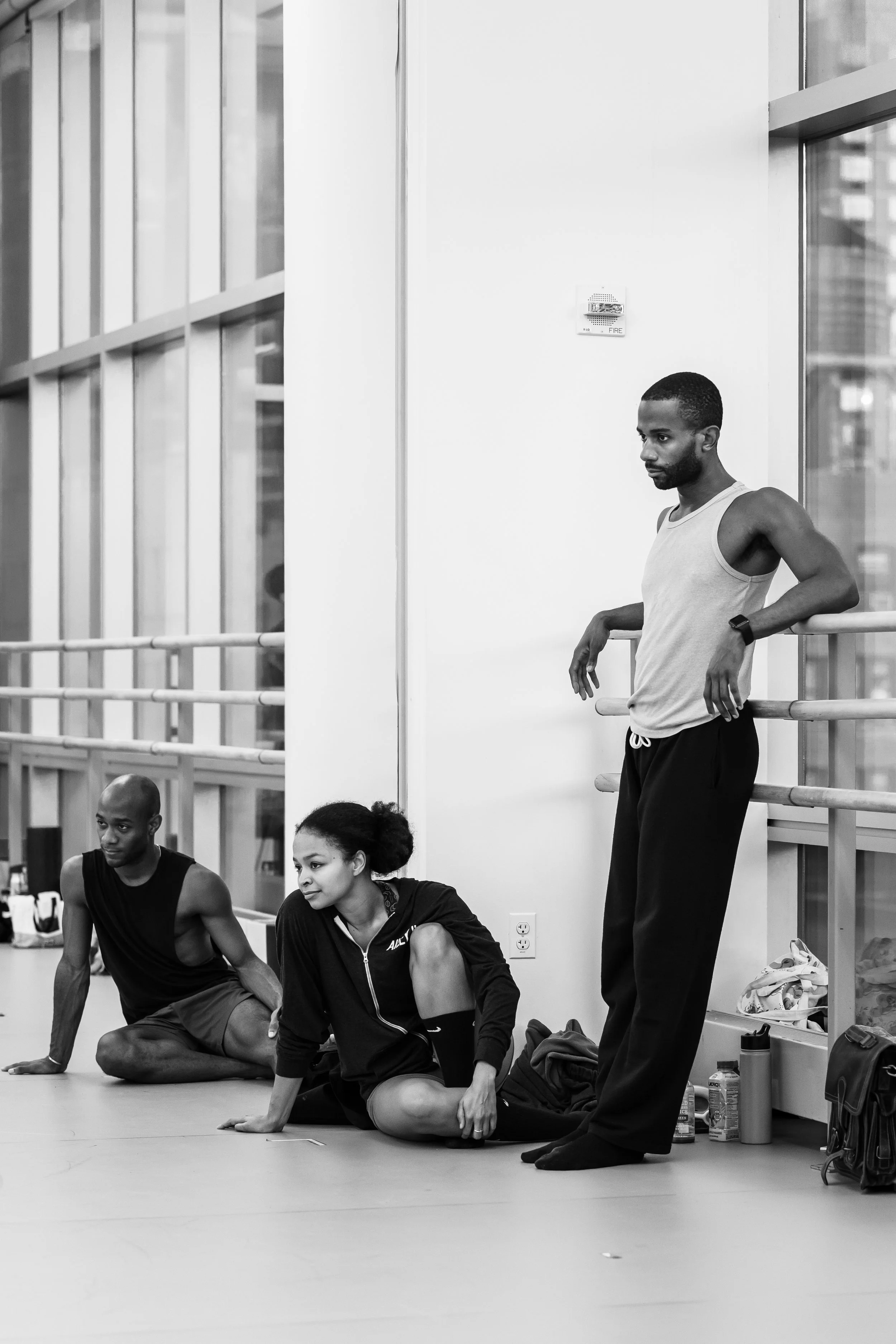 Group of people in workout clothes sitting and stretching on the floor of a fitness studio, with a man standing and leaning against a ballet barre.