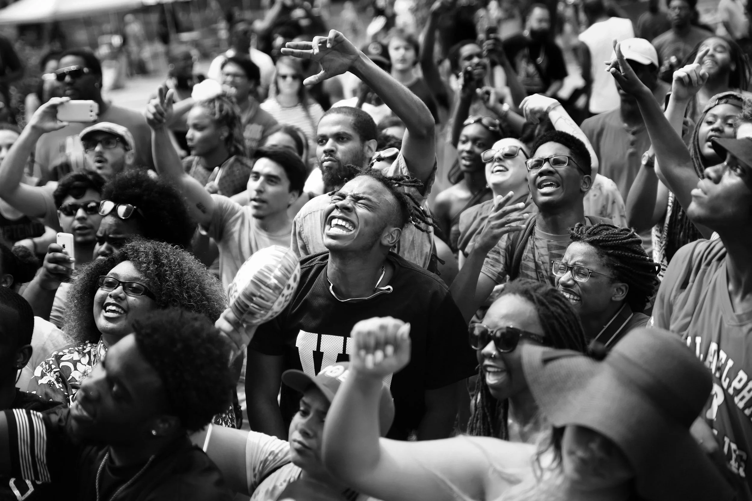 A large crowd of people gathered outdoors, some making hand gestures and taking photos, suggesting participation in a protest or rally.