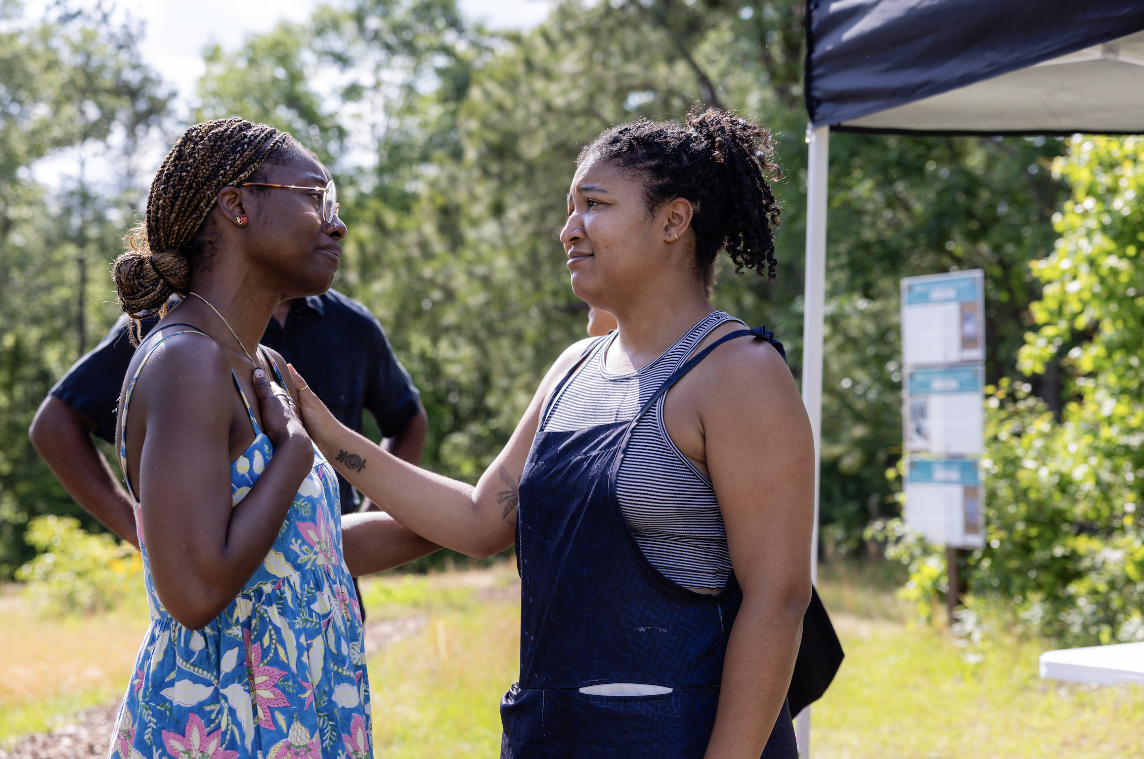 Two women facing each other, emotional moment outdoors in a park, one holding her hand over her heart, the other reaching out gently, surrounded by trees and sunlight.