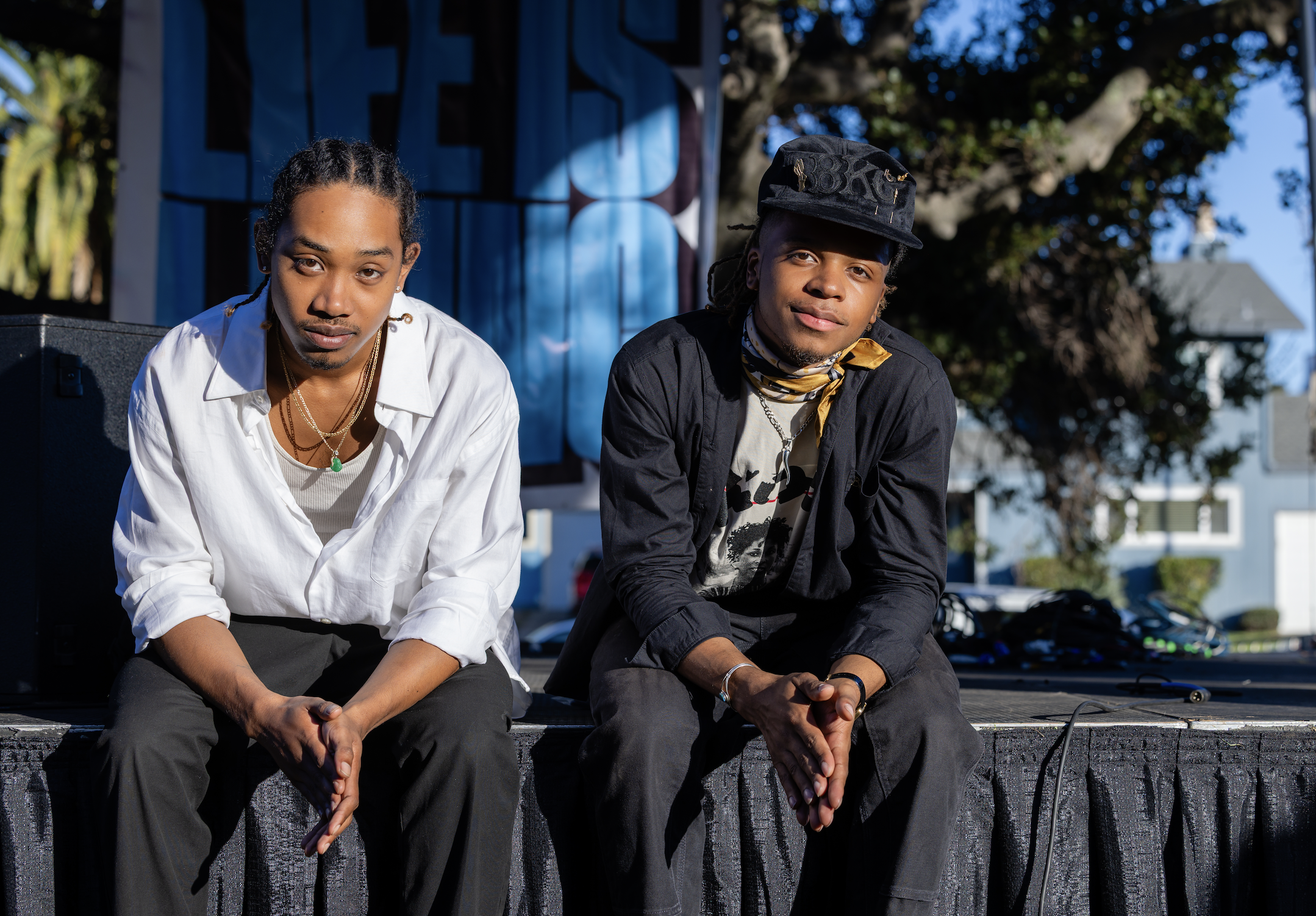 Two young men sitting on a raised platform outdoors in daylight. One is wearing a white shirt with jewelry, and the other is dressed in black with a black cap and a scarf around his neck. Both are looking at the camera with relaxed expressions.