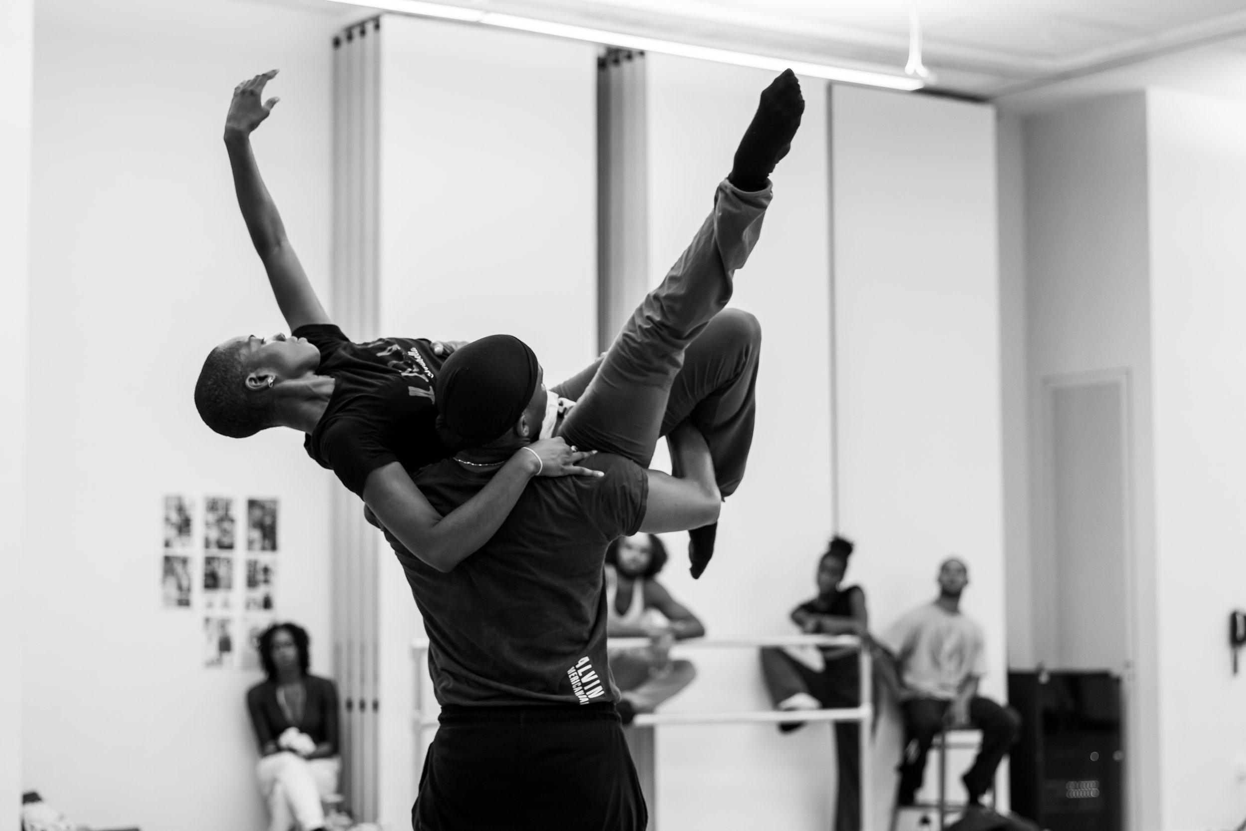 Two individuals practicing Brazilian jiu-jitsu in a martial arts class, with one person executing a submission hold on the other, in a training room with spectators watching in the background.