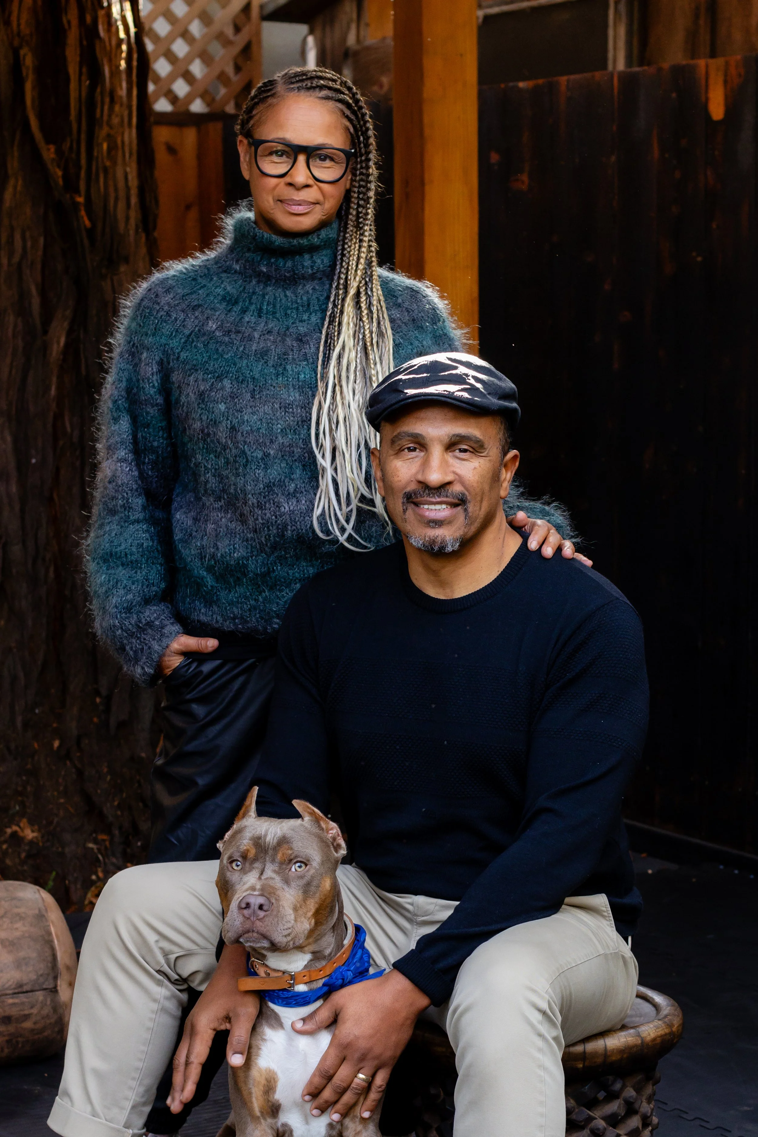 A woman with braided hair and glasses, wearing a cozy sweater, stands behind a seated man with a dog on his lap. The man is wearing a black shirt, beige pants, and a cap. The dog has a brown and white coat and is wearing a blue bandana. They are outd