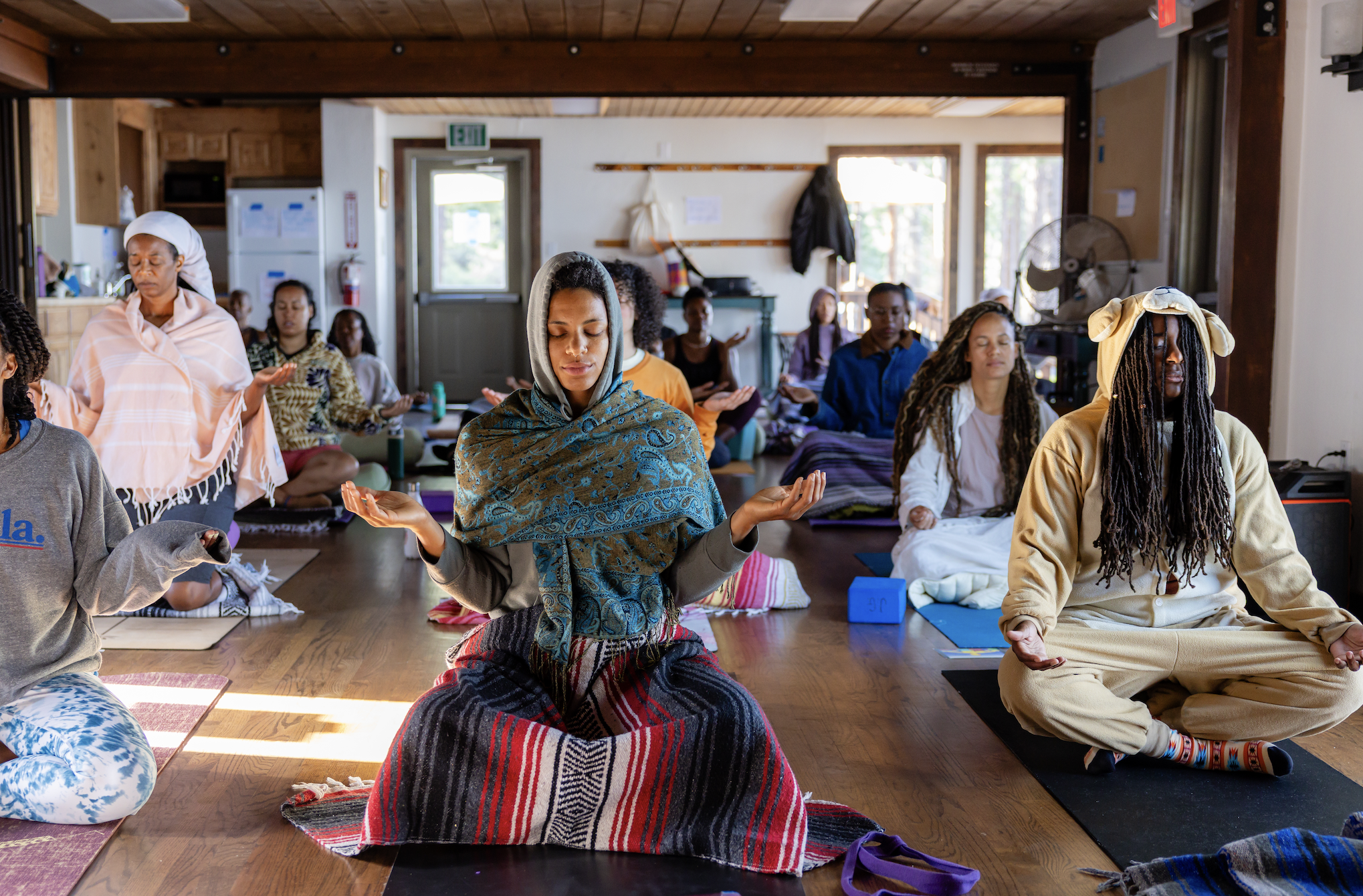 A group of diverse people practicing meditation indoors, sitting cross-legged on mats with closed eyes and peaceful expressions, with sunlight streaming through windows in the background.