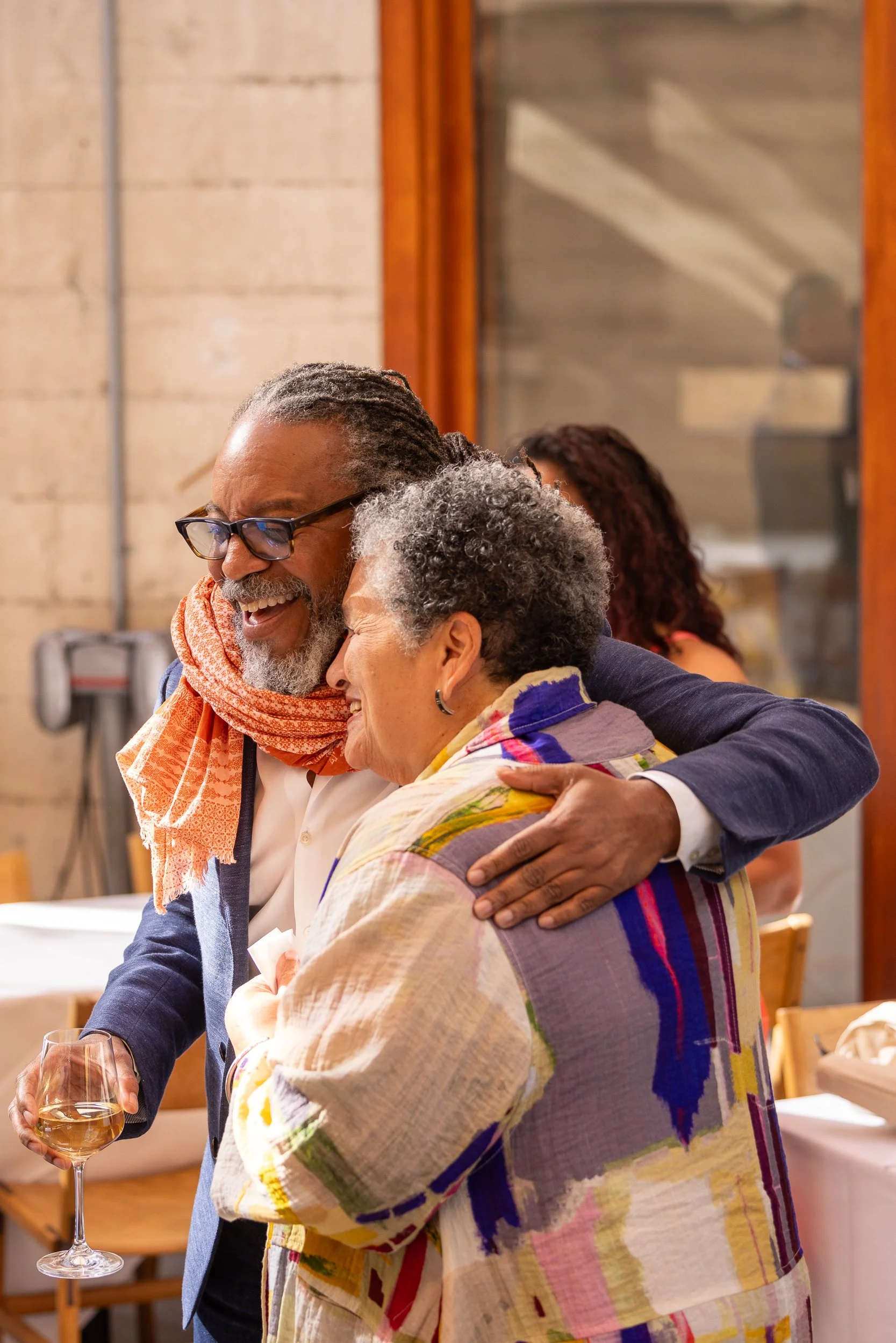 A man and an older woman are hugging, both smiling and happy, indoors at a social gathering, with the man holding a glass of wine.