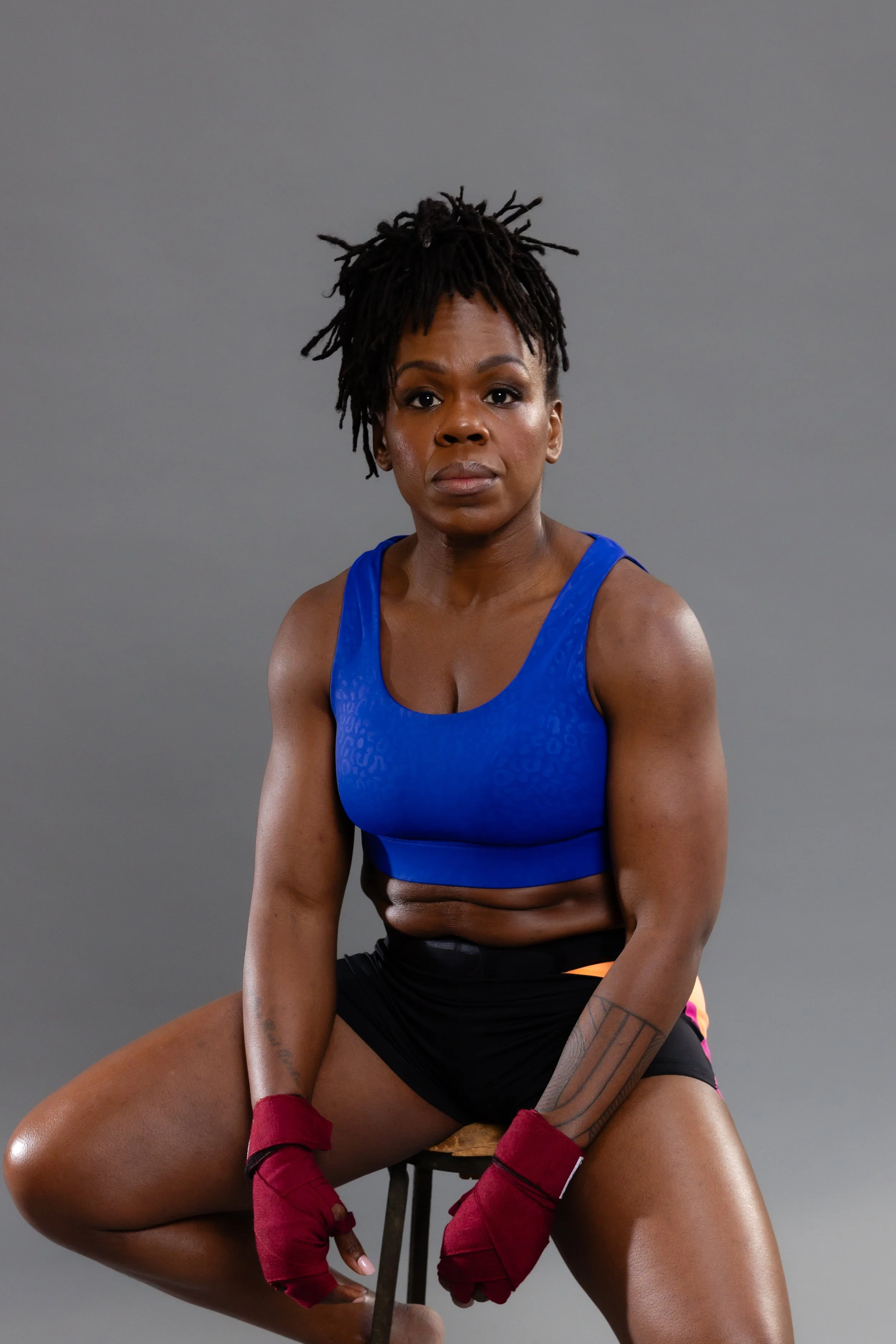 A woman in a blue sports top and black shorts is sitting on a stool against a plain gray background, wearing red boxing gloves, with her hair in dreadlocks, looking directly at the camera.
