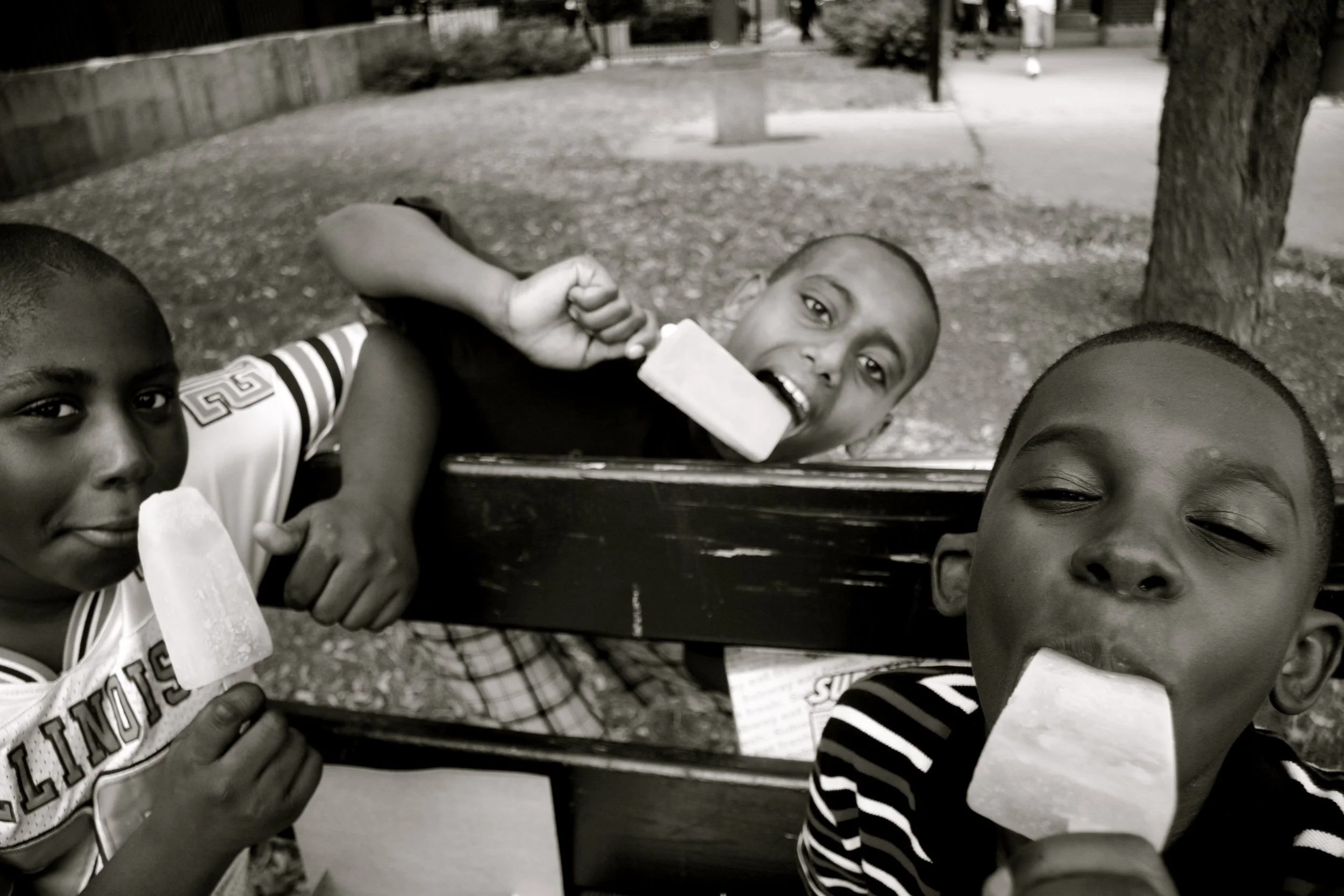 Three young boys enjoying ice pops while sitting on a park bench, with trees and a sidewalk in the background.