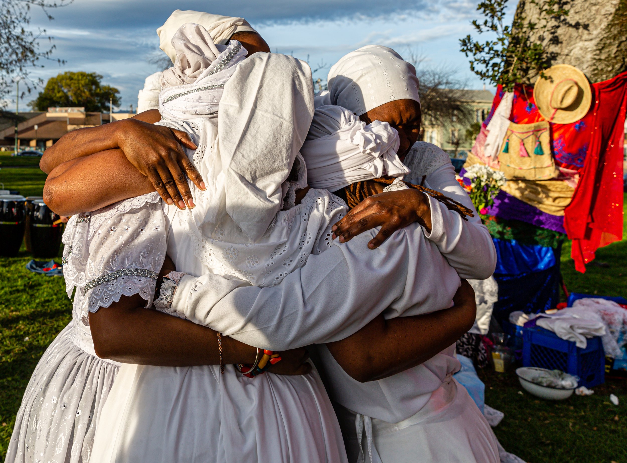 Two women dressed in white, hugging outdoors during daytime, with vibrant clothing and accessories on display nearby.