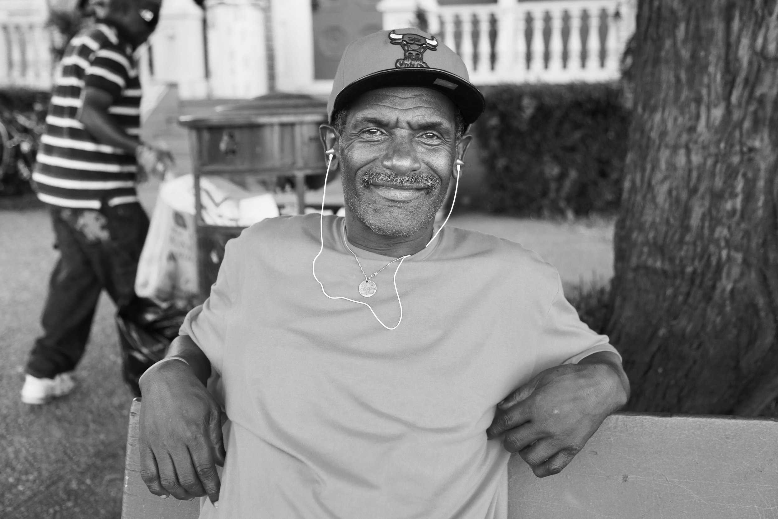 Black and white photo of an older man sitting outside, smiling slightly, wearing a baseball cap with the Chicago Bulls logo, earphones, and a T-shirt, with a young person in the background walking away.