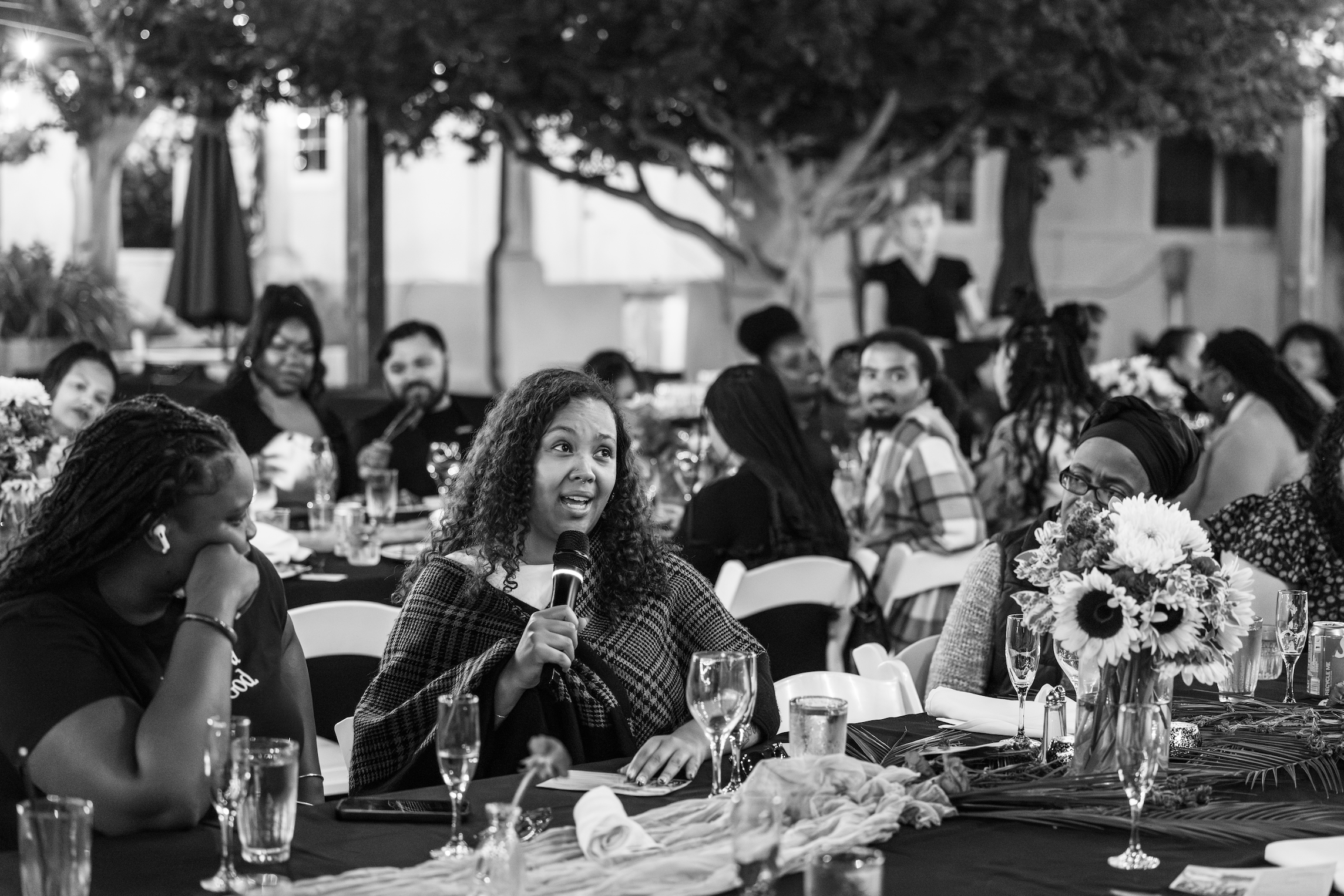 A woman with curly hair holding a microphone and speaking at a formal gathering. She is seated at a table decorated with a bouquet of flowers, wine glasses, and tableware. Other guests are visible in the background, sitting at similar tables in an ou