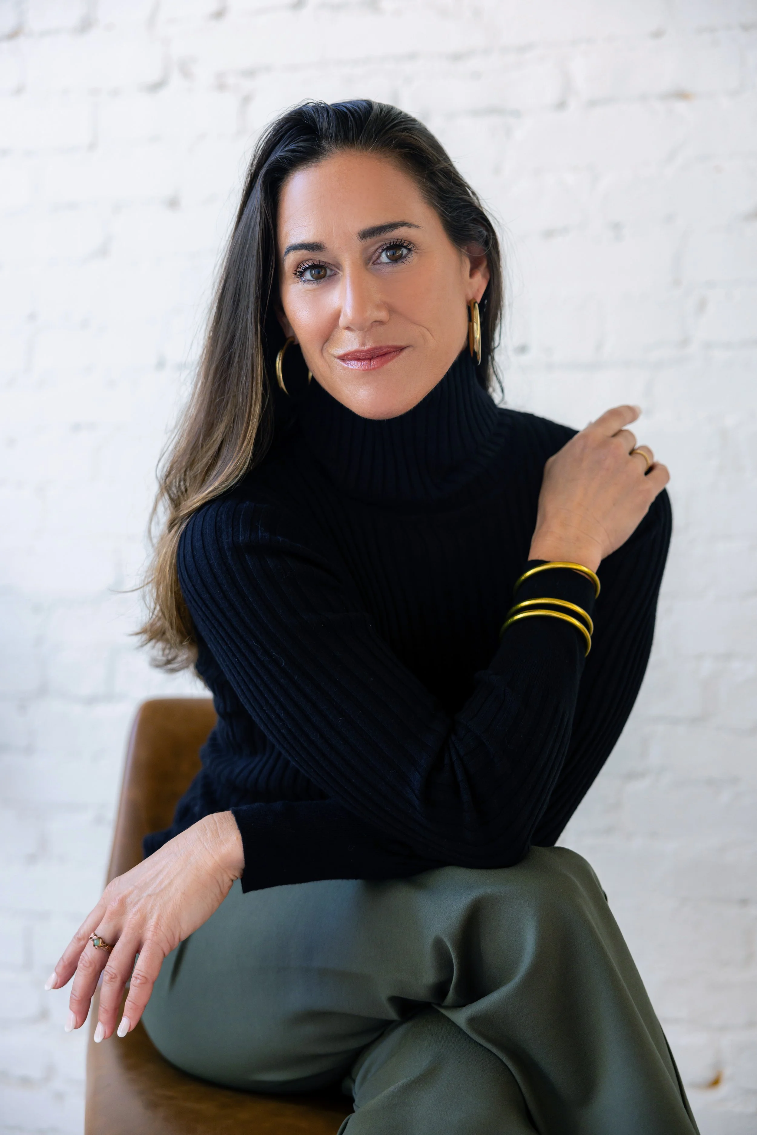 A woman with long dark hair, wearing a black turtleneck sweater and gold jewelry, sitting on a chair against a white brick wall.