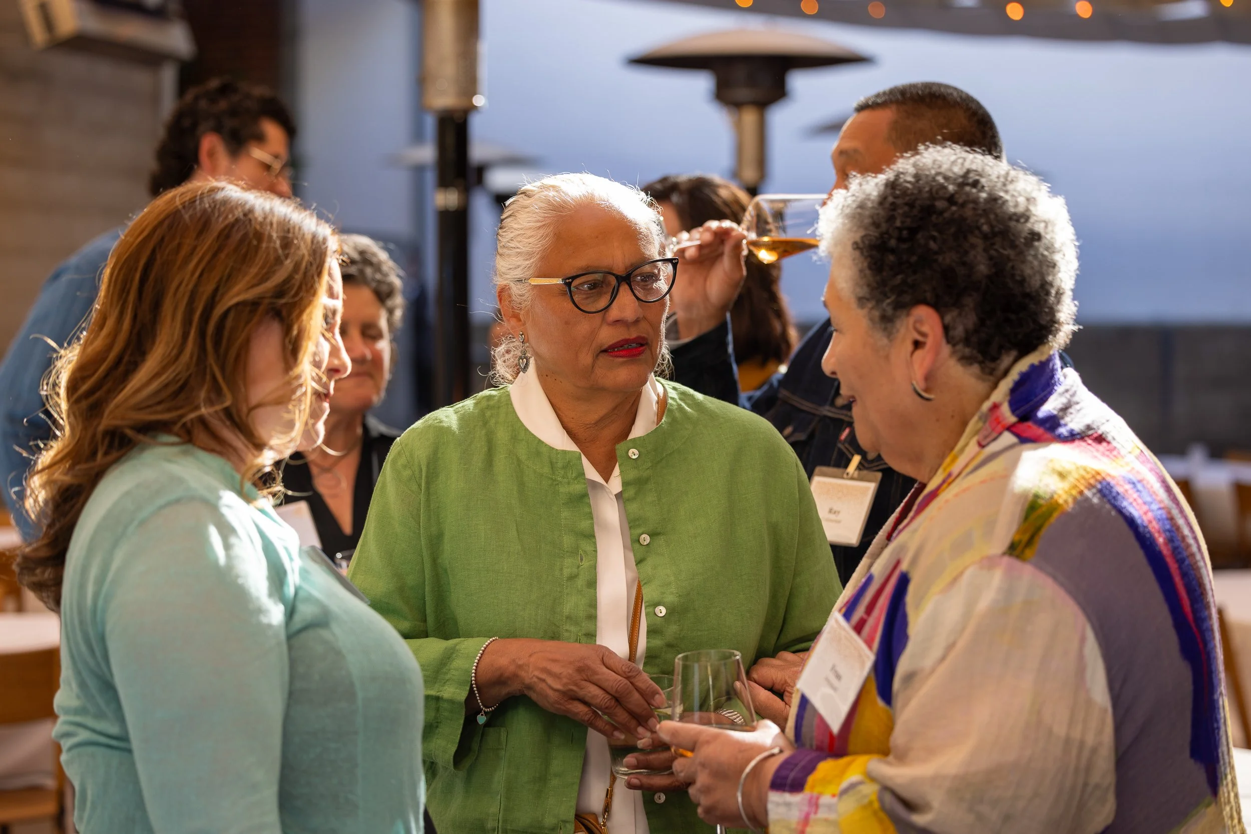 Group of women at social event, one woman in green sweater holding a wine glass, engaging in conversation.