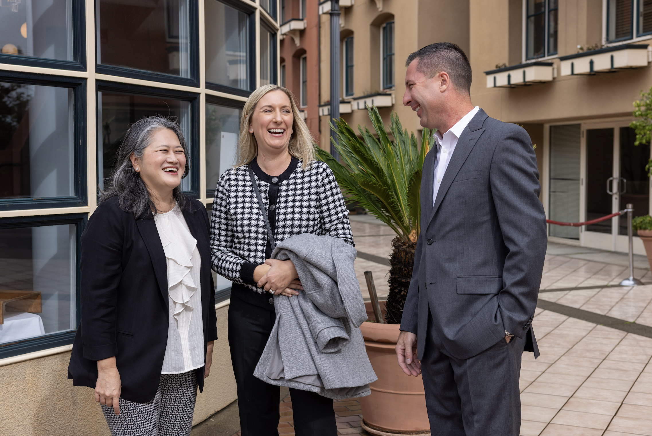 Three people, one man and two women, are standing outside a building, smiling and talking. The woman on the left has gray hair and is wearing a black blazer. The woman in the middle has blonde hair and is wearing a black and white houndstooth jacket.