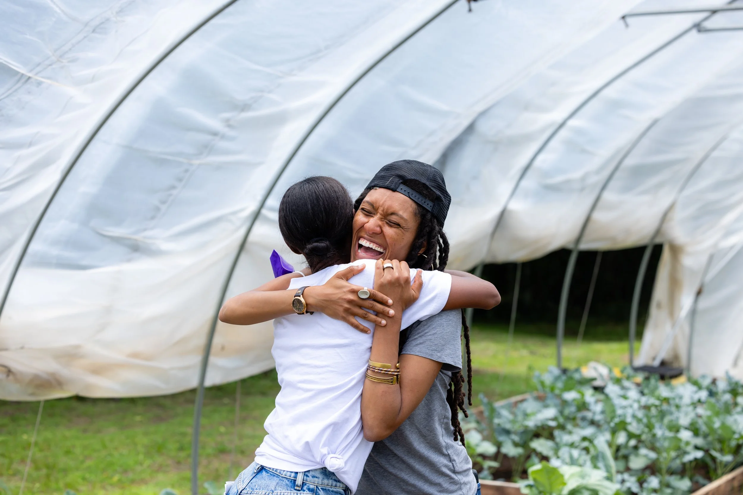Two women are hugging happily in a greenhouse or garden, with greenery and plants around them.