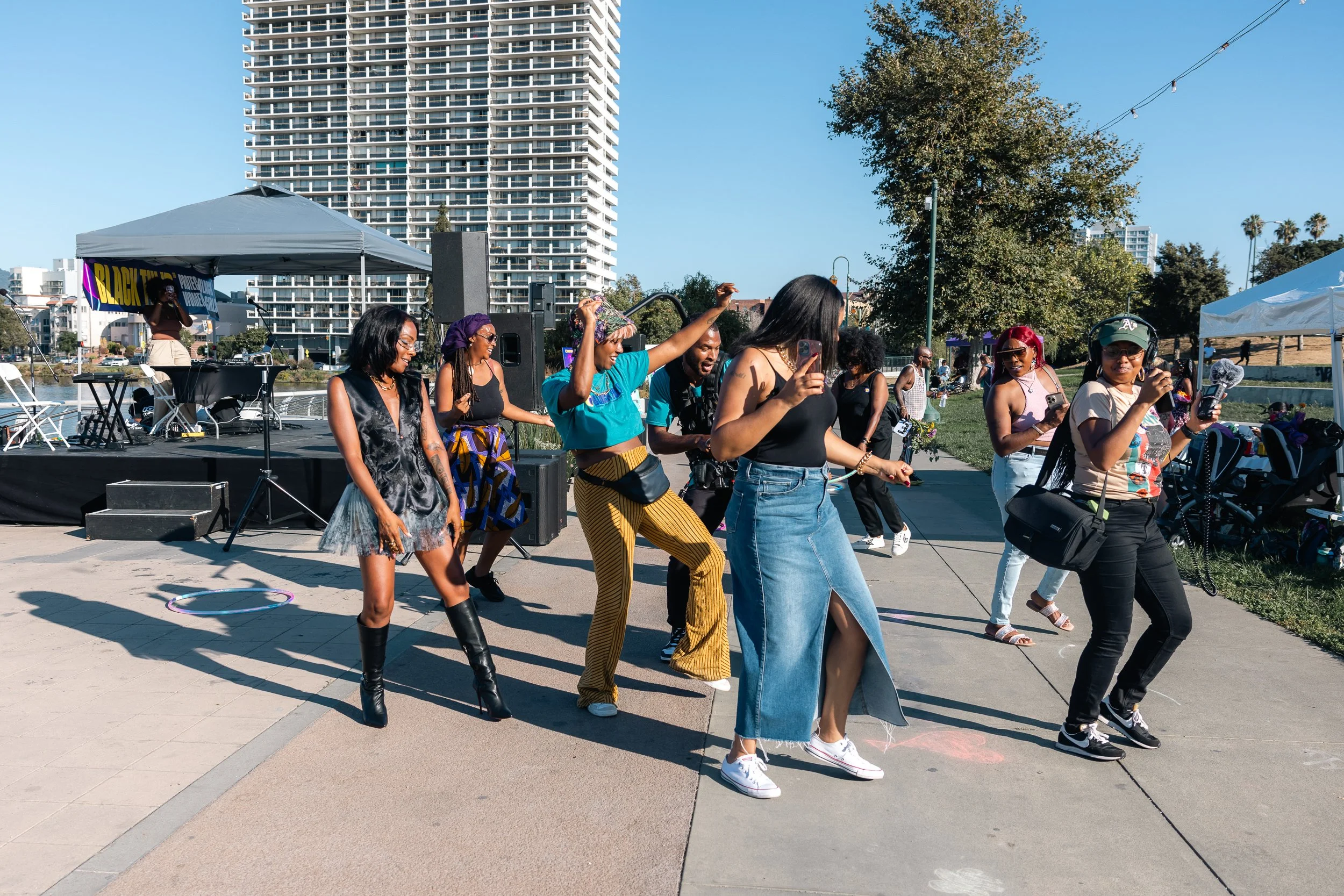 People dancing outdoors near a stage with a speaker, in a park area by the water, with high-rise buildings and trees in the background on a sunny day.