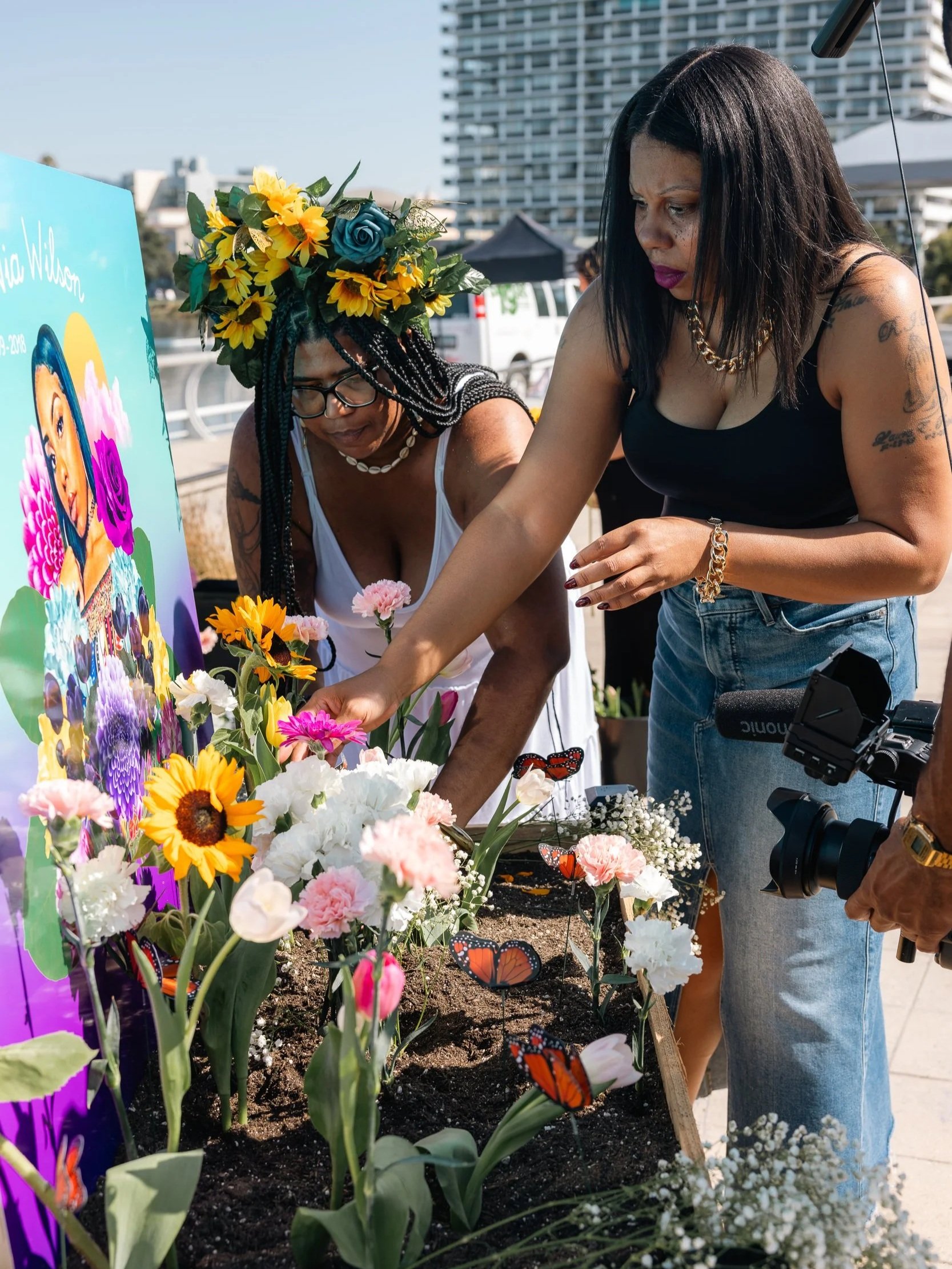 Two women are planting flowers in a garden bed. One woman has long black hair, tattoos, and is wearing a black top and blue jeans. The other woman has braids with a large floral headpiece and is wearing glasses and a white dress. There are colorful flowers and butterfly decorations in the soil, with city buildings in the background.
