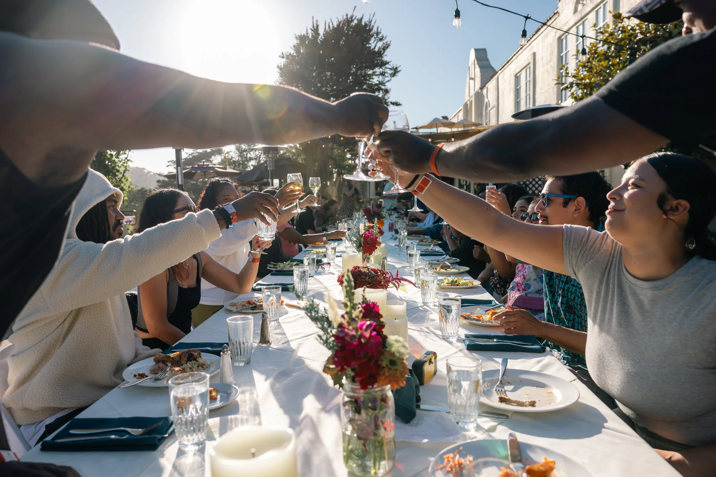 People sitting at a long outdoor dining table, raising glasses for a toast, with sunlight shining from behind, surrounded by flowers and surrounded by buildings and string lights.