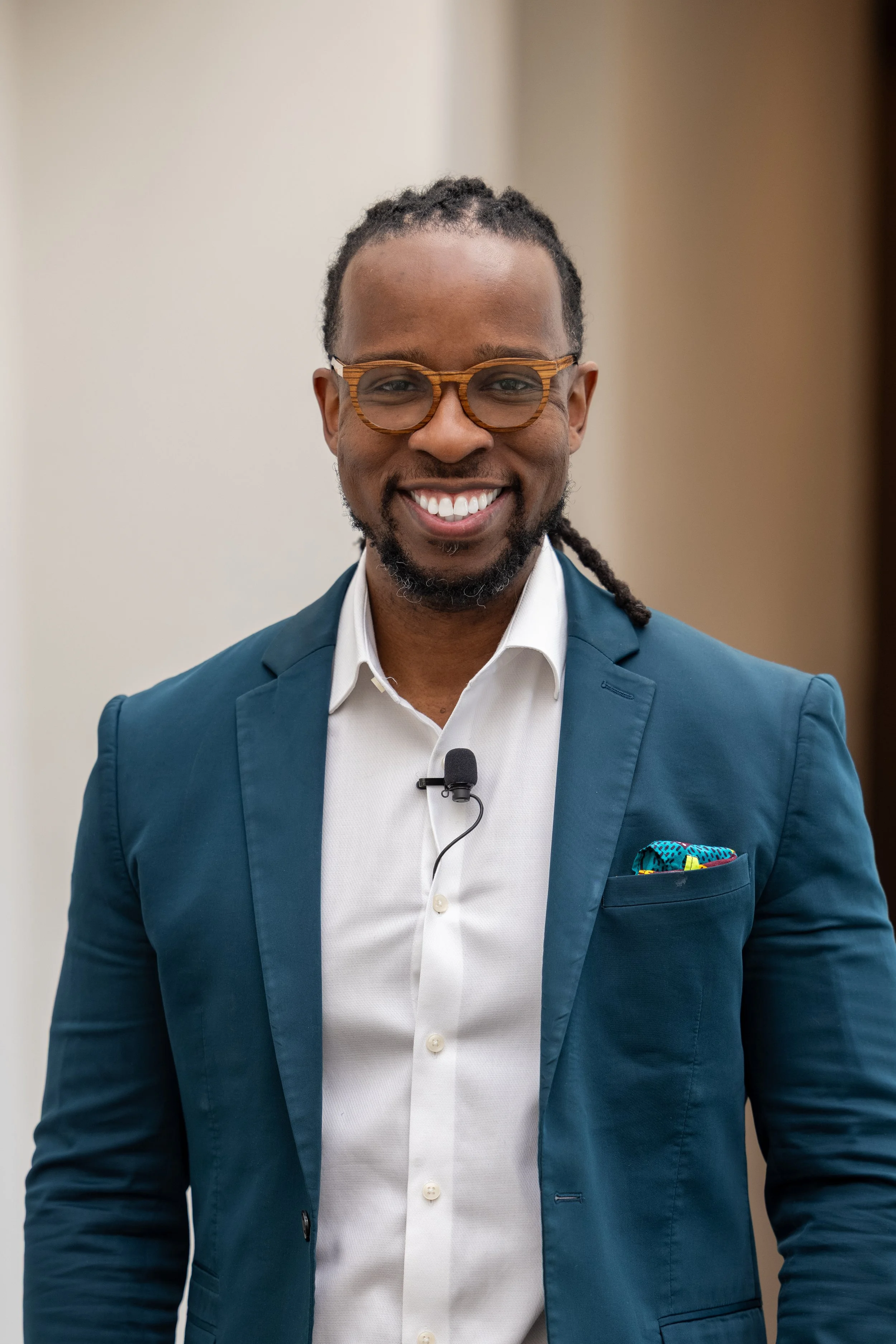 A smiling man wearing glasses and a blue blazer stands indoors, dressed in a white shirt with a microphone and a colorful pocket square in his blazer pocket.