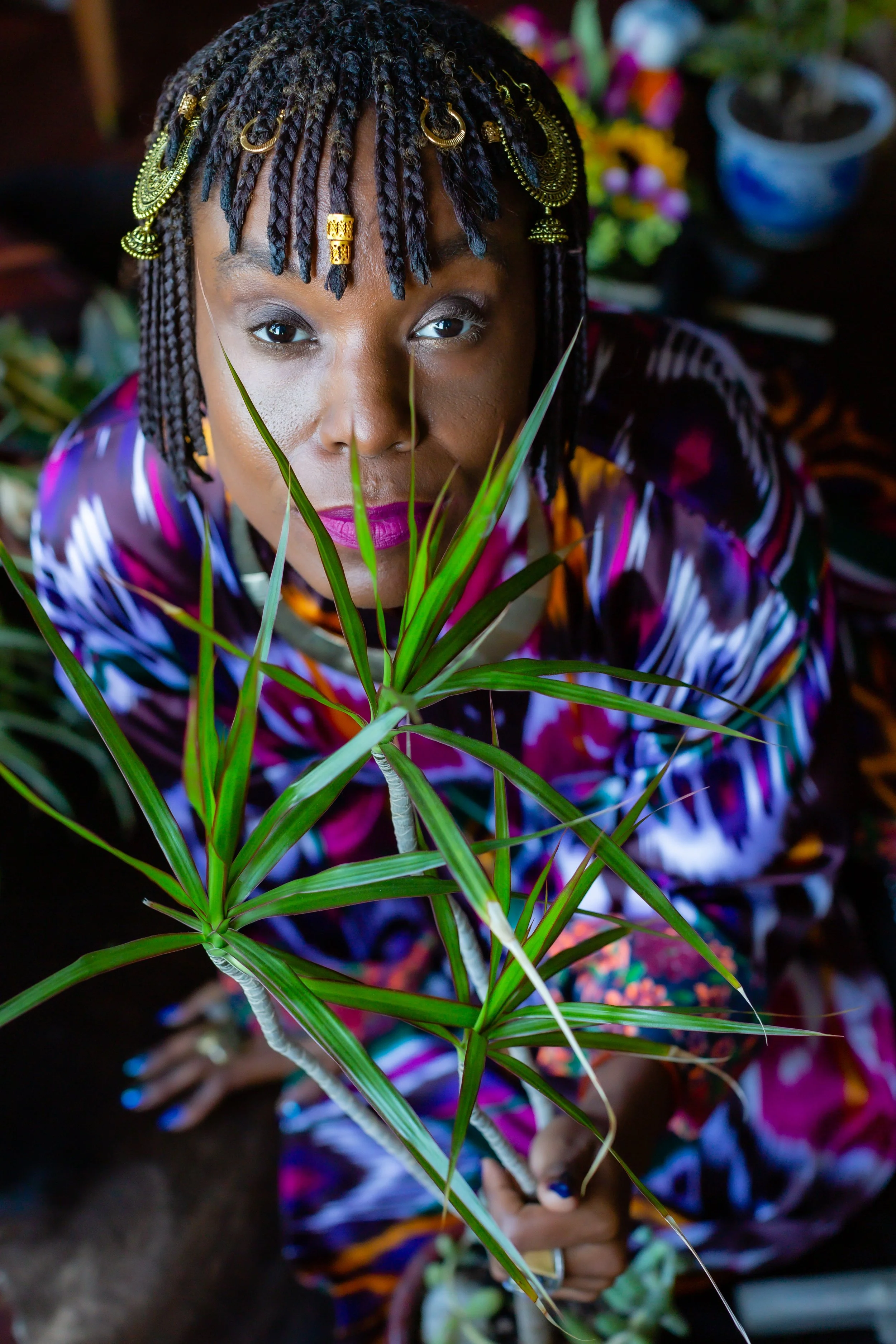 A woman with braided hair adorned with gold jewelry, holding a green plant in front of her face, with colorful flowers in the background.