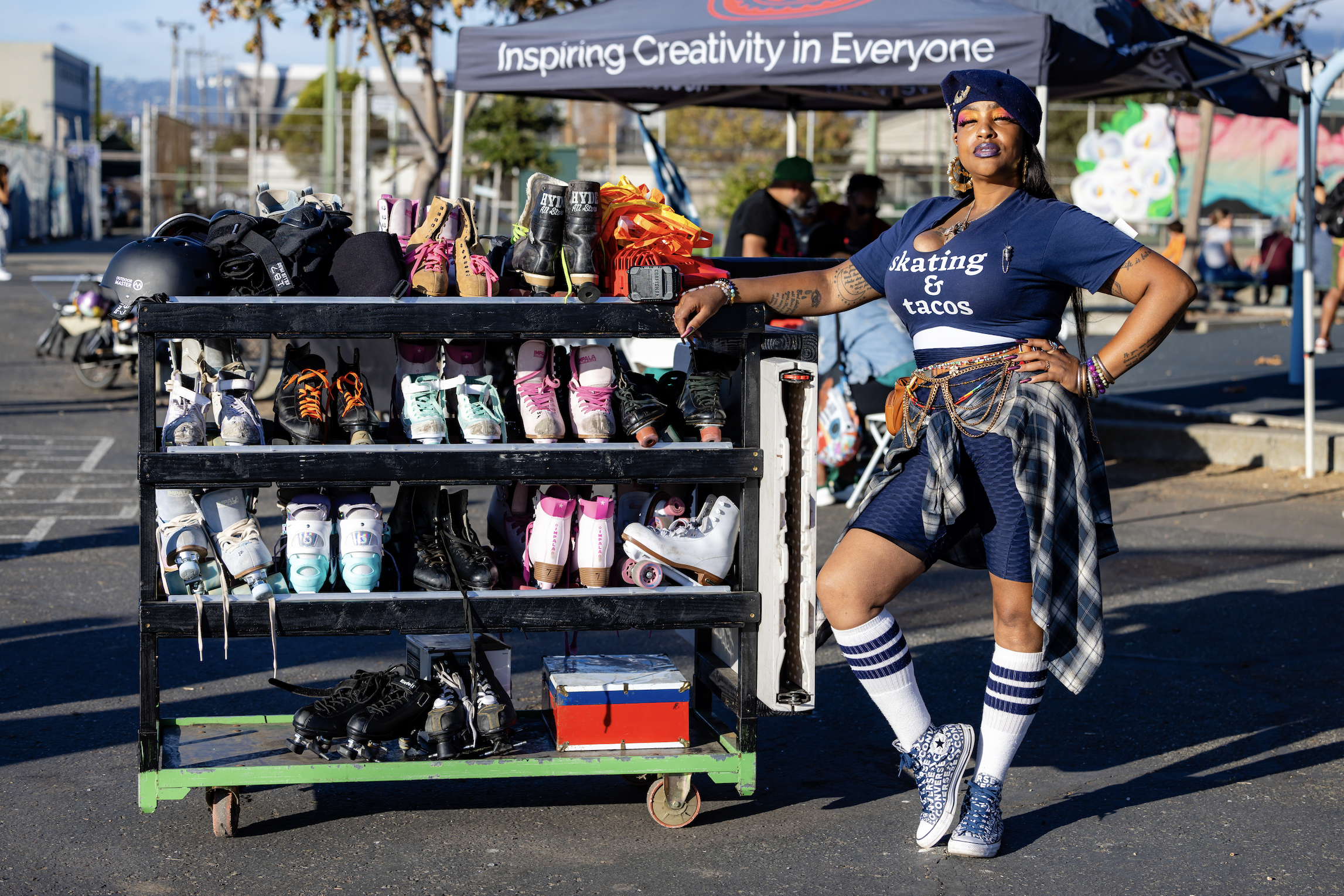 A woman with dark skin, wearing a blue beret, a blue T-shirt that says "skating & tacos," plaid shirt tied around her waist, striped knee-high socks, and Converse sneakers, stands beside a skate rental cart with various roller skates and helmets at a