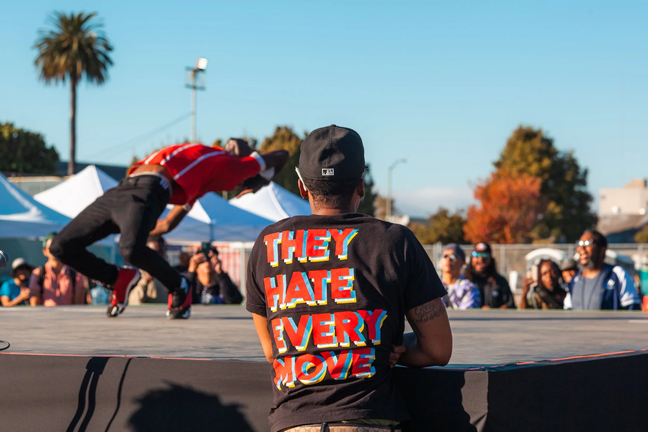 Person standing with back to camera, wearing a black cap and a black t-shirt with colorful text overlay that says "THEY HATE EVERY" during outdoor event, with other people and tents in the background.
