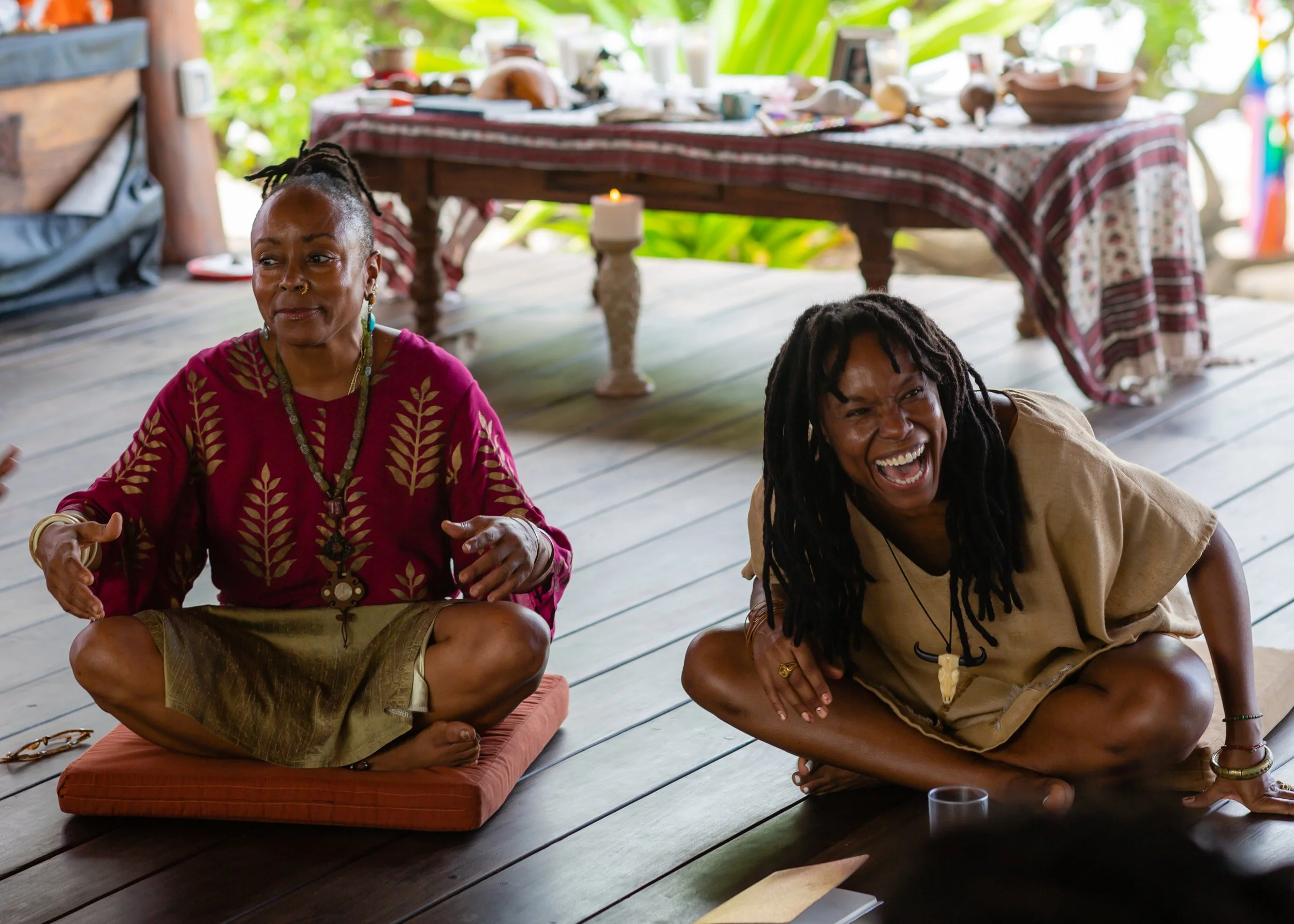 Two women sitting on a wooden floor, one with dreadlocks and the other with a bun, smiling and laughing indoors with a table set with various objects behind them.