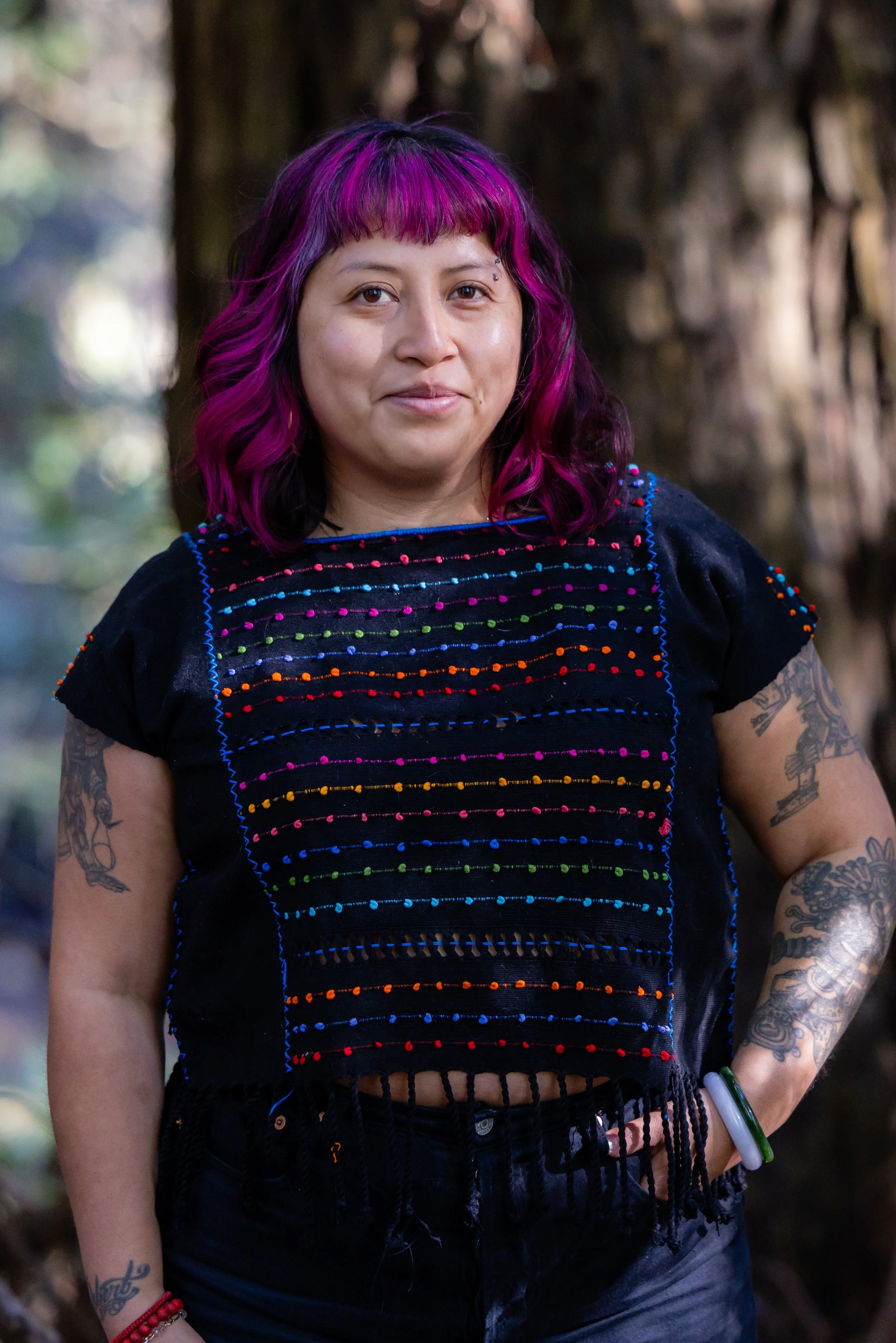 A woman with vibrant pink and purple hair standing outdoors in a forest. She has tattoos on her arms and is wearing a black top with colorful embroidered patterns and fringes. She is smiling subtly.