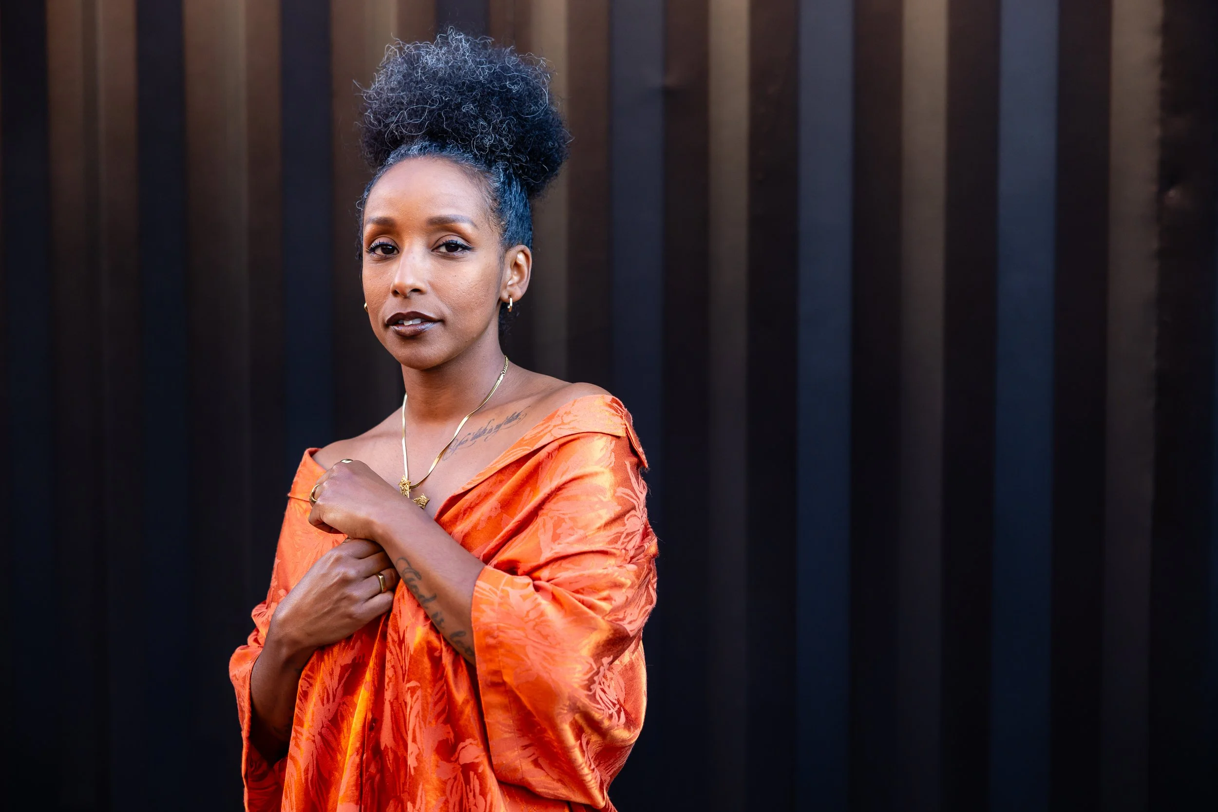 A woman with curly black hair styled in an updo, wearing an orange patterned dress and gold jewelry, standing against a dark, vertical striped background.