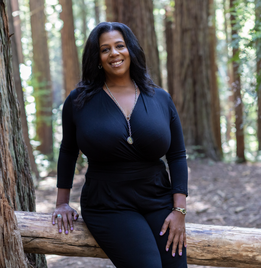 A woman in a black outfit sitting on a fallen log in a forest with tall trees and green foliage.