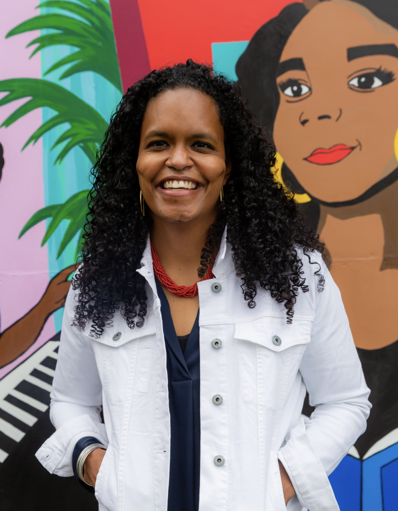 A woman with curly black hair smiling and wearing a white jacket, red necklace, and gold earrings, standing in front of a colorful mural with a woman with dark hair and a yellow earring.