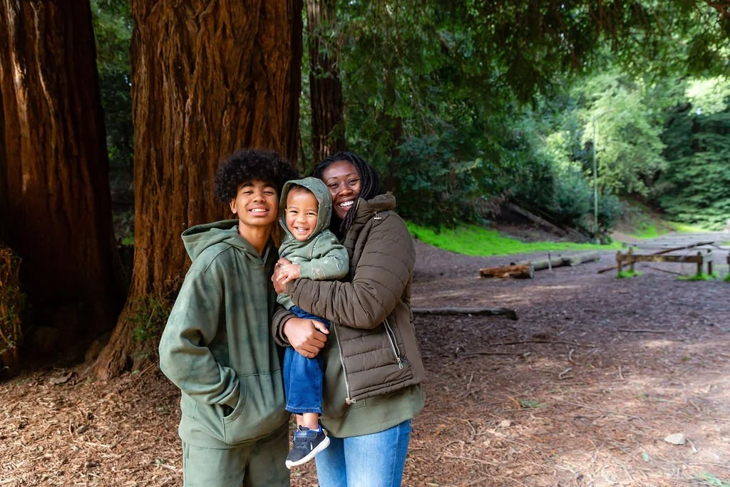 Three people, a woman and two children, smiling and hugging in a forest with large trees and green foliage.