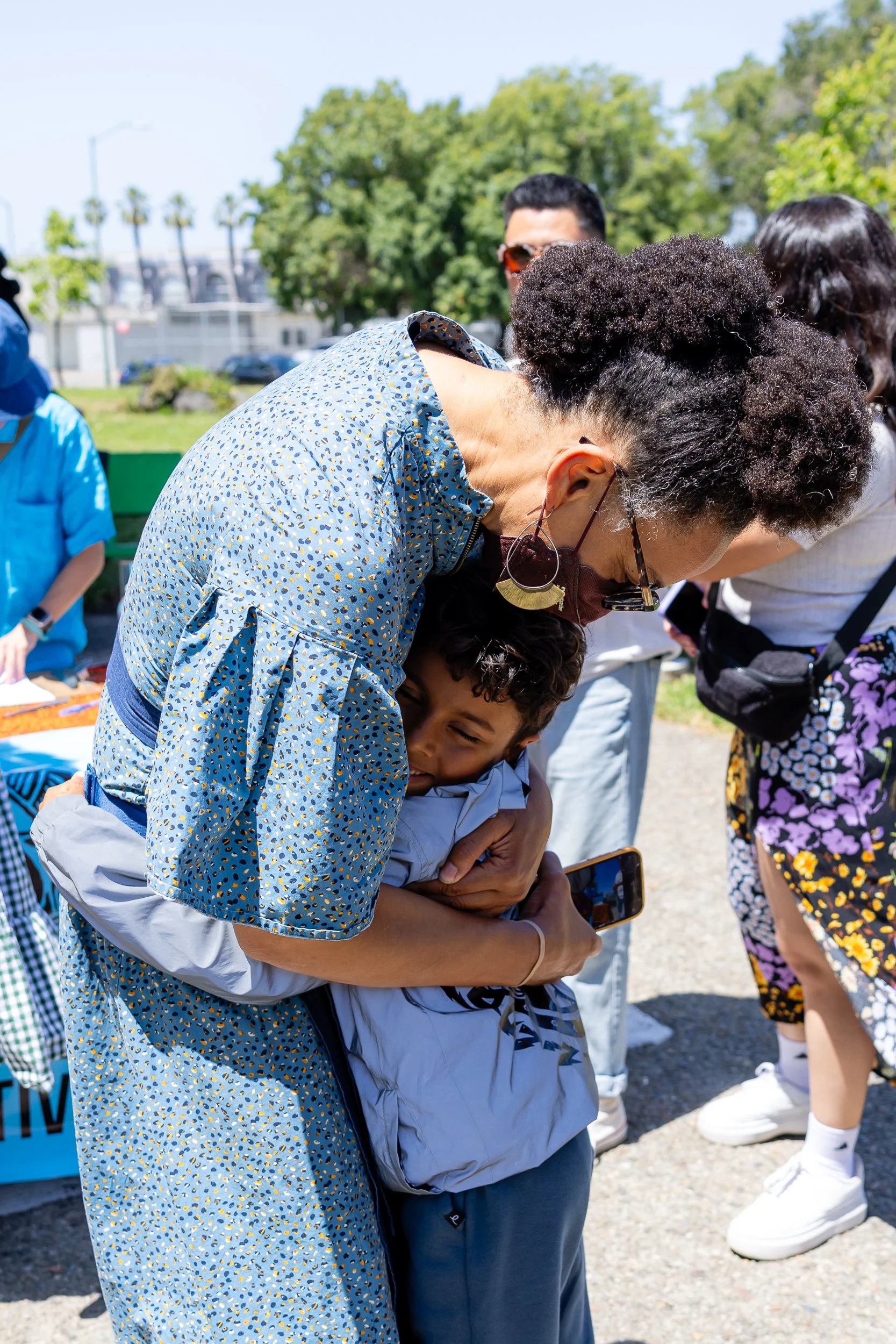 A woman and a young boy hugging each other outdoors on a sunny day, with people and trees in the background.