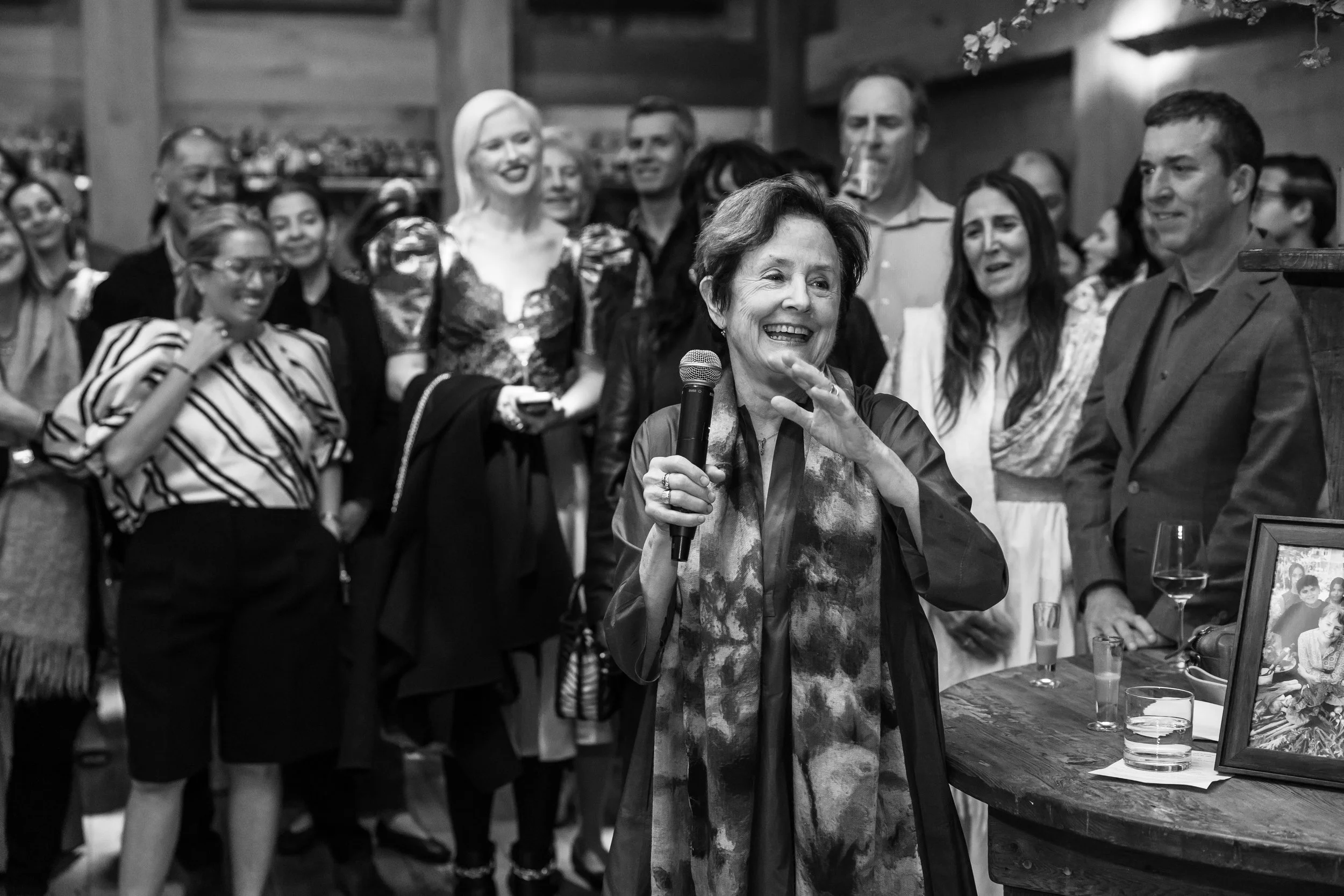 A woman speaking into a microphone at a gathering, with a group of smiling people standing behind her, some holding drinks. The scene appears to be in a rustic indoor setting with wooden walls.