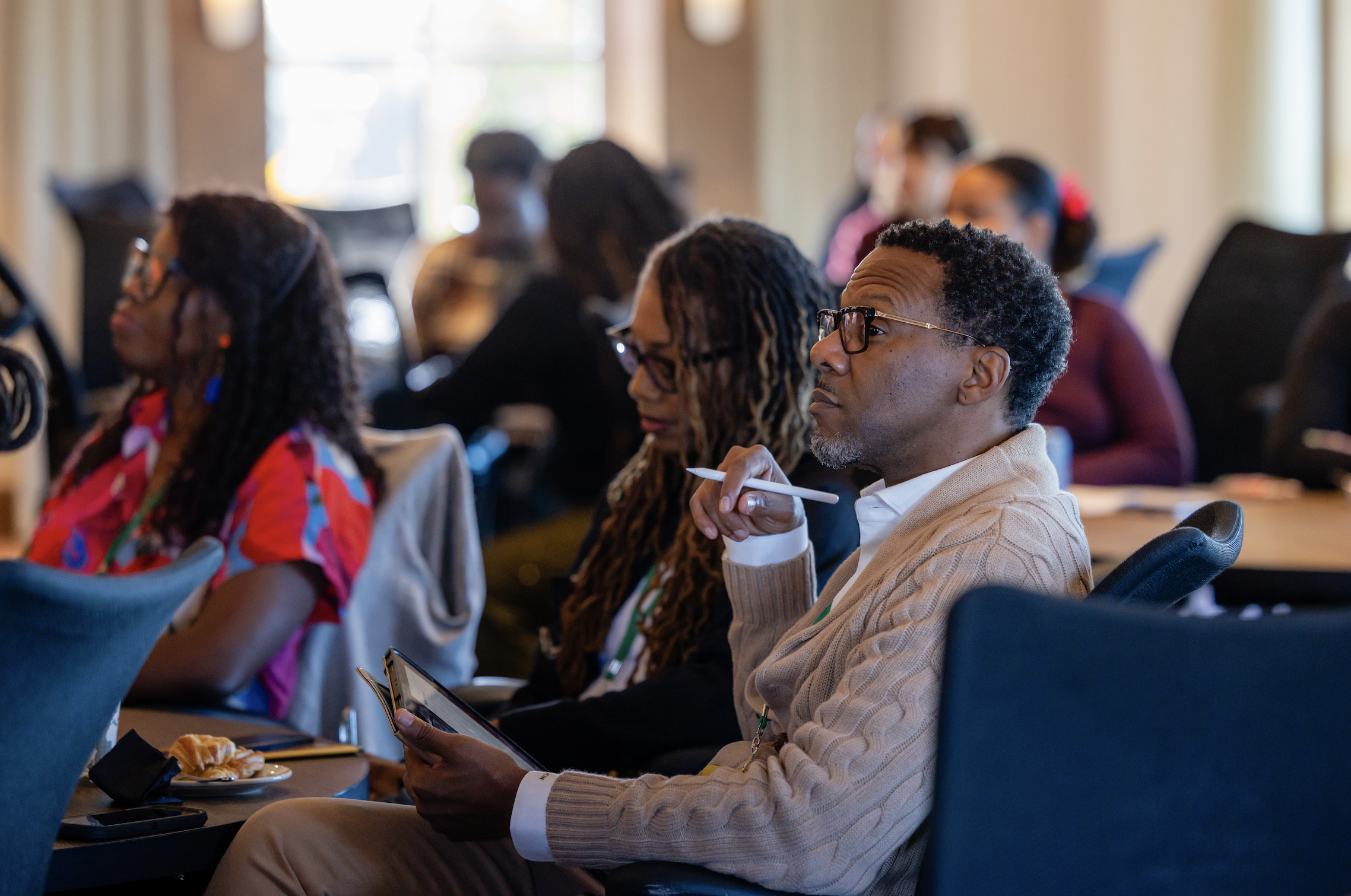 People attending a conference or seminar in a well-lit room, sitting at tables, some taking notes, with a focus on a man with glasses holding a pen and a woman with dreadlocks reading a book or pamphlet.