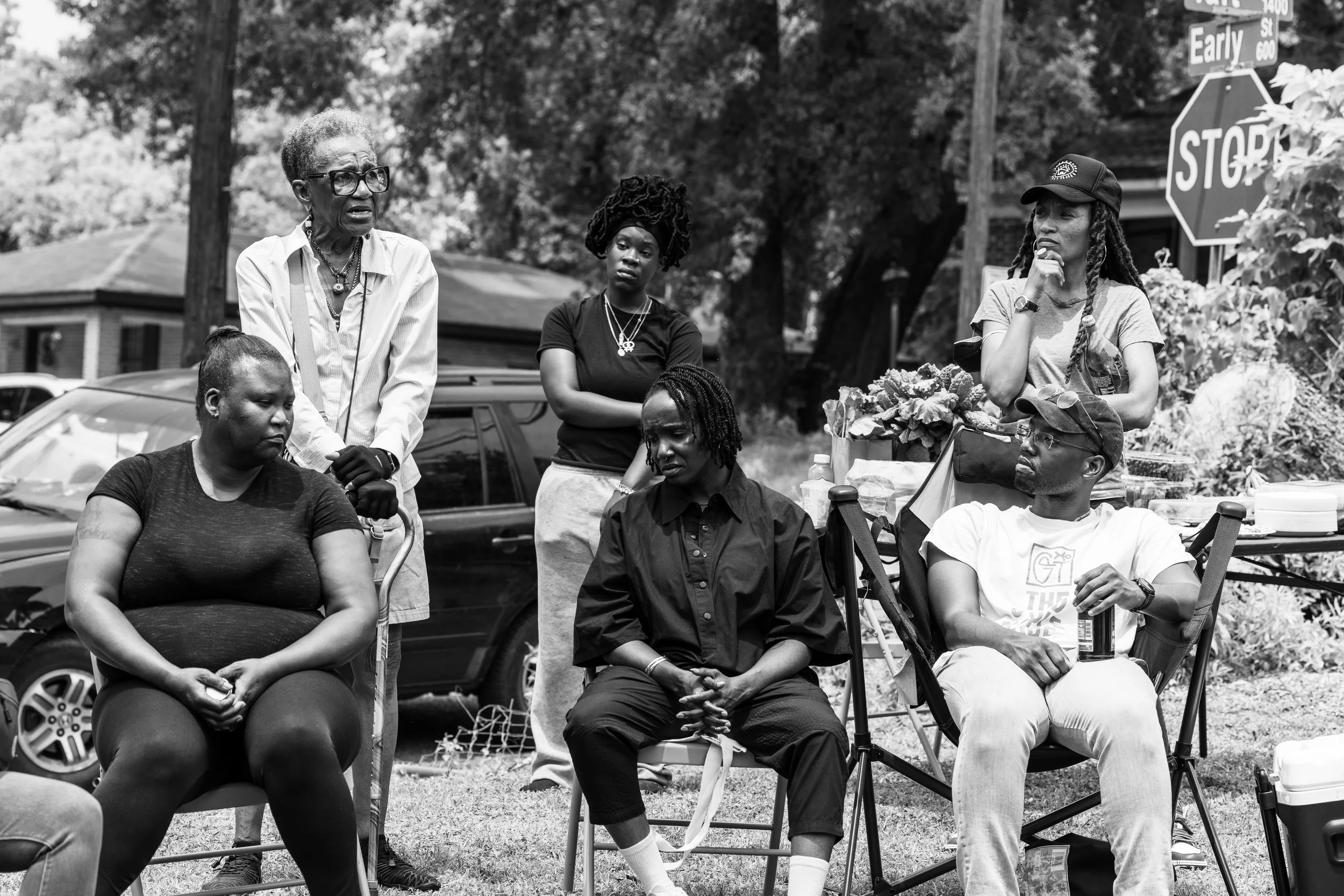A group of women sitting and standing outdoors, engaged in a discussion, with trees and cars in the background.