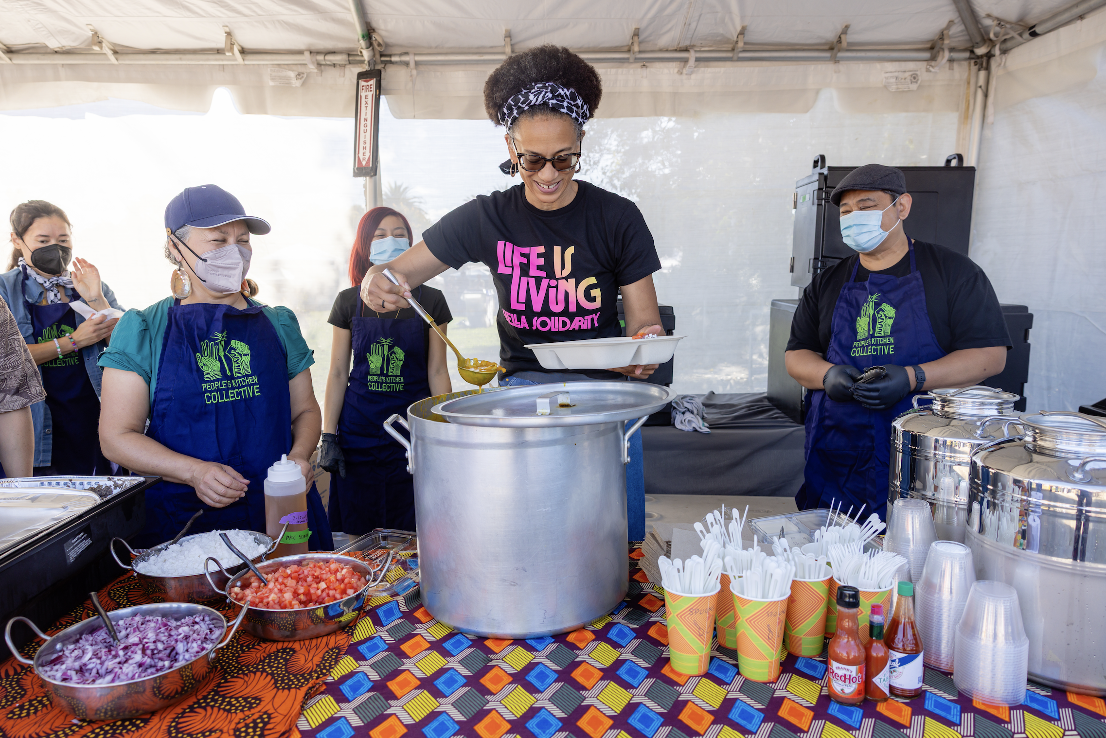 A group of diverse women and a man, all wearing face masks and aprons, serving food at a community event under a tent. The table has bowls of chopped vegetables, condiments, and disposable cups with utensils.