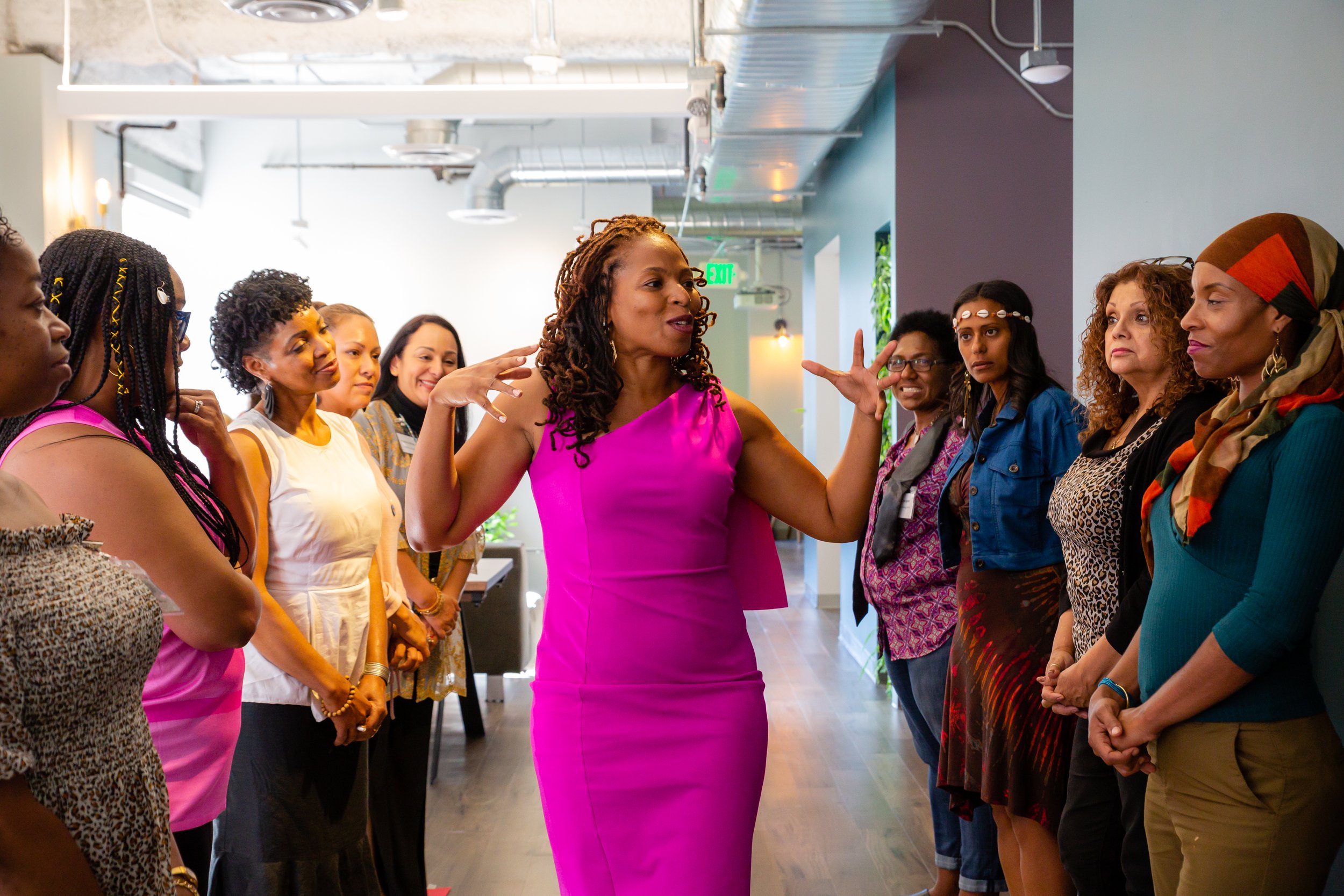 A woman in a pink dress appears to be speaking to a group of women standing in a line indoors, with exposed ductwork and a modern interior background. Event photography.