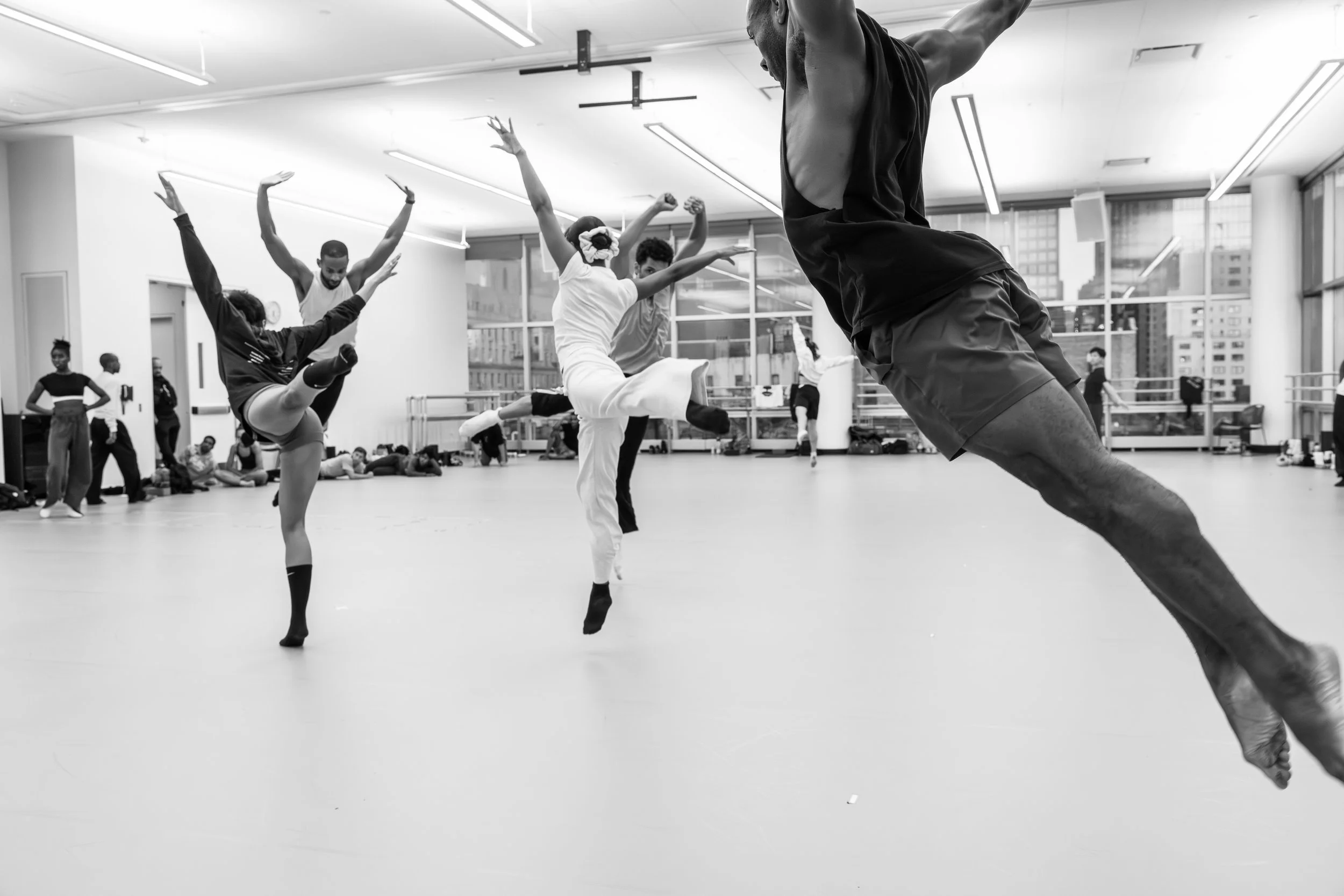 Ballet dancers practicing in a dance studio with large windows, some dancers are on one leg with arms raised, others are on the floor stretching or resting, and a dance instructor is leading the class, all in black and white.