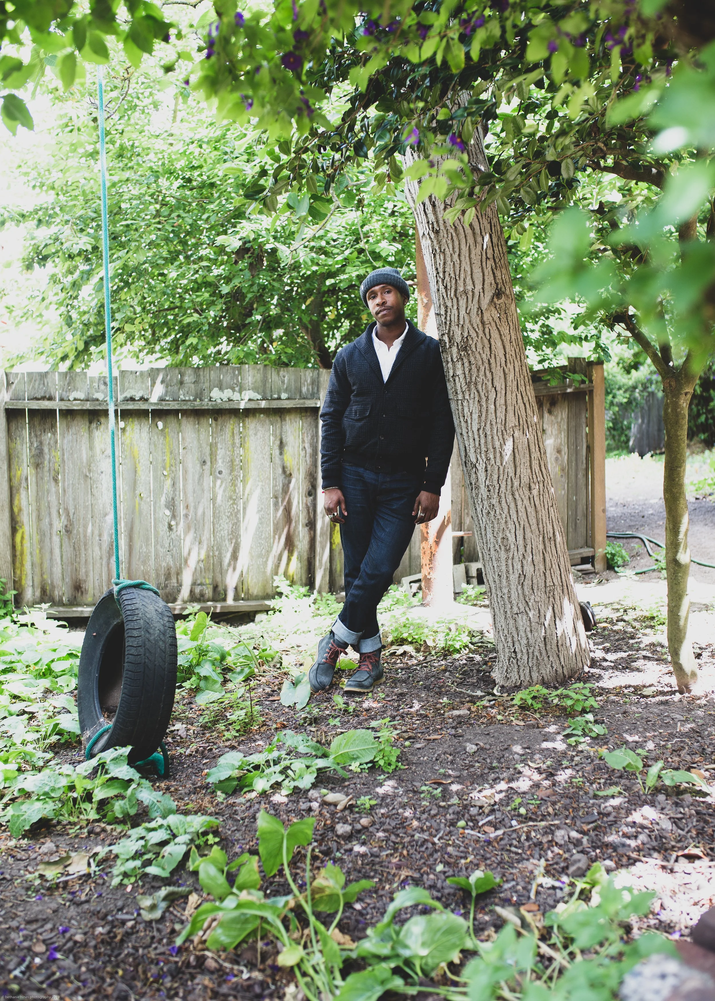 A young man wearing a black beanie, white shirt, and black jacket leaning against a large tree in a backyard, with a wooden fence, green foliage, and a tire in the foreground.