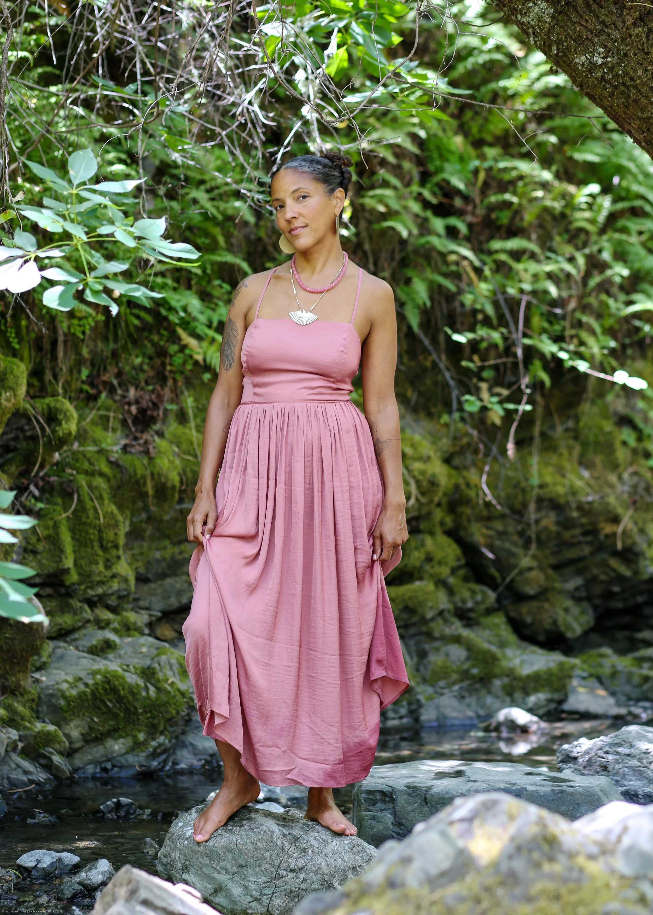 A woman in a pink dress standing barefoot on rocks in a stream surrounded by green foliage.