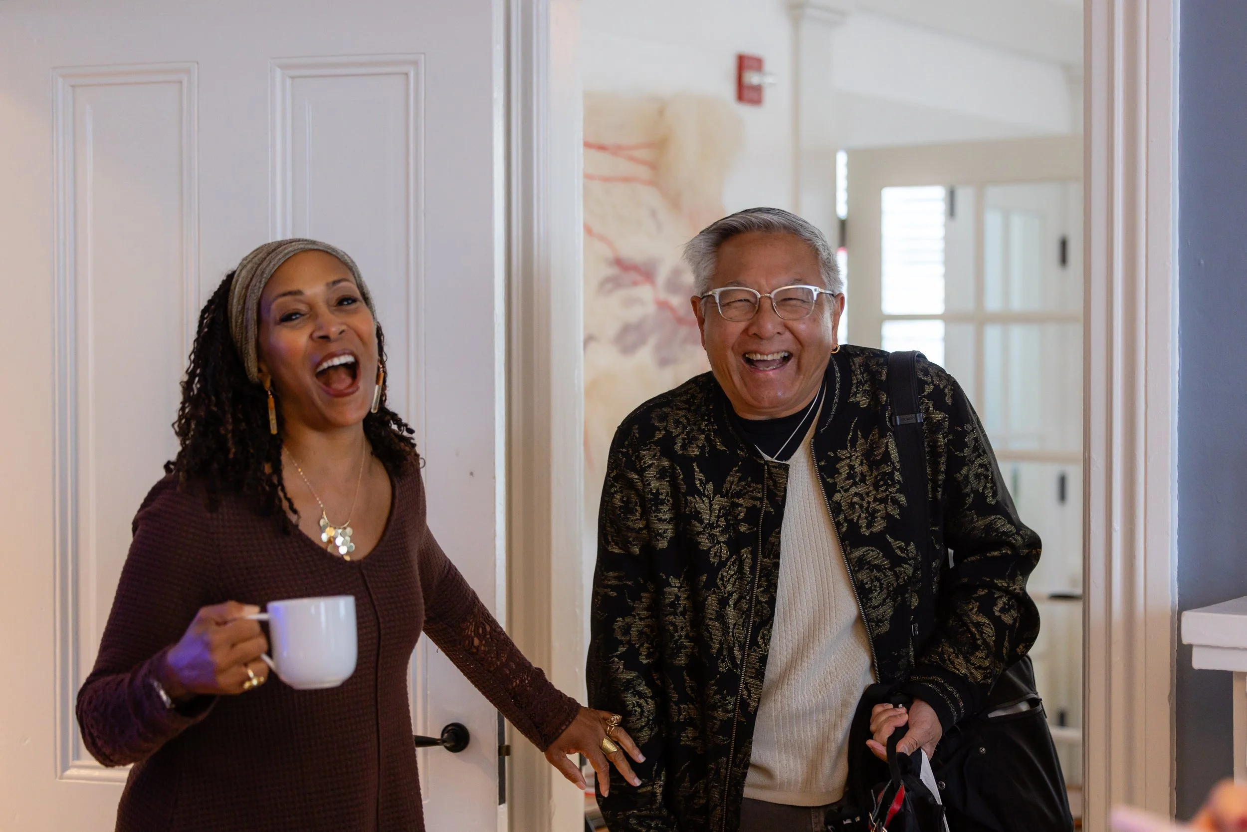 Two women and one man smiling and laughing in an indoor setting. The woman on the left holds a white coffee mug, and their hands are intertwined. They appear to be happy and enjoying each other's company.