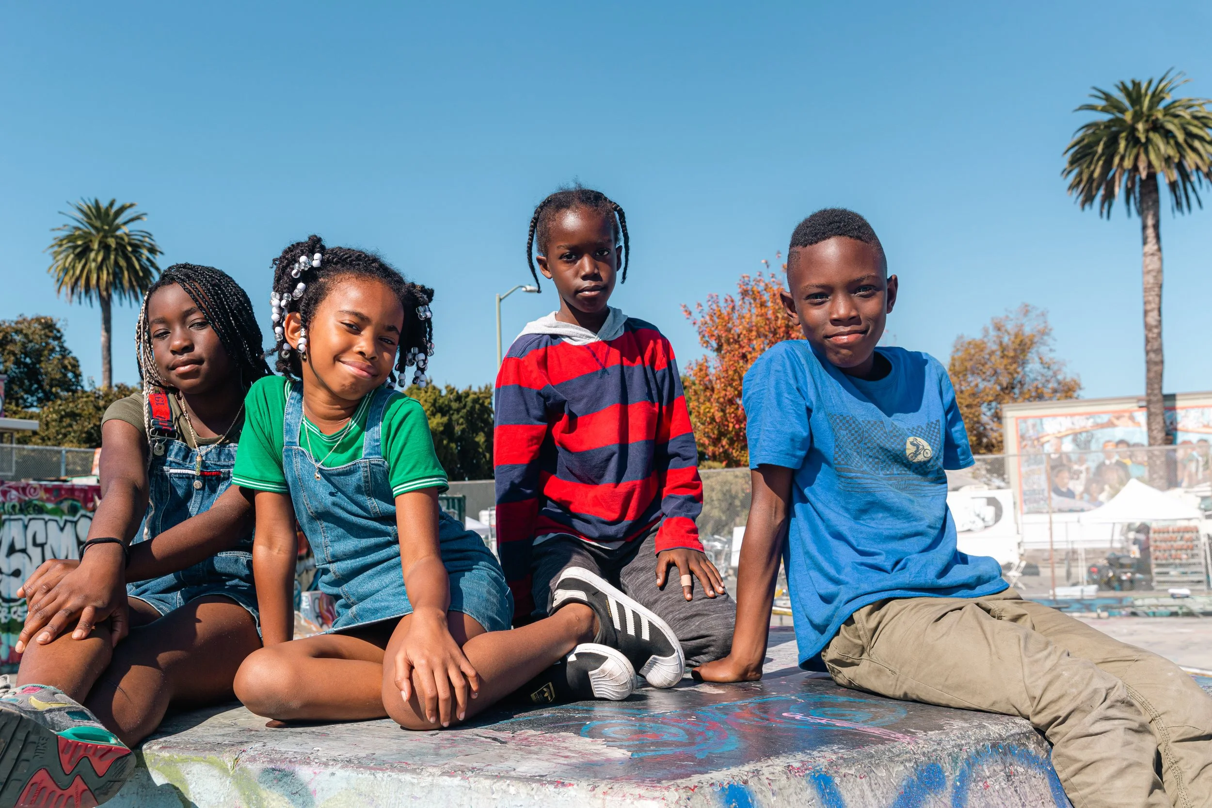 Four children sitting on a graffiti-covered ledge outdoors on a sunny day, with palm trees and a blue sky in the background.