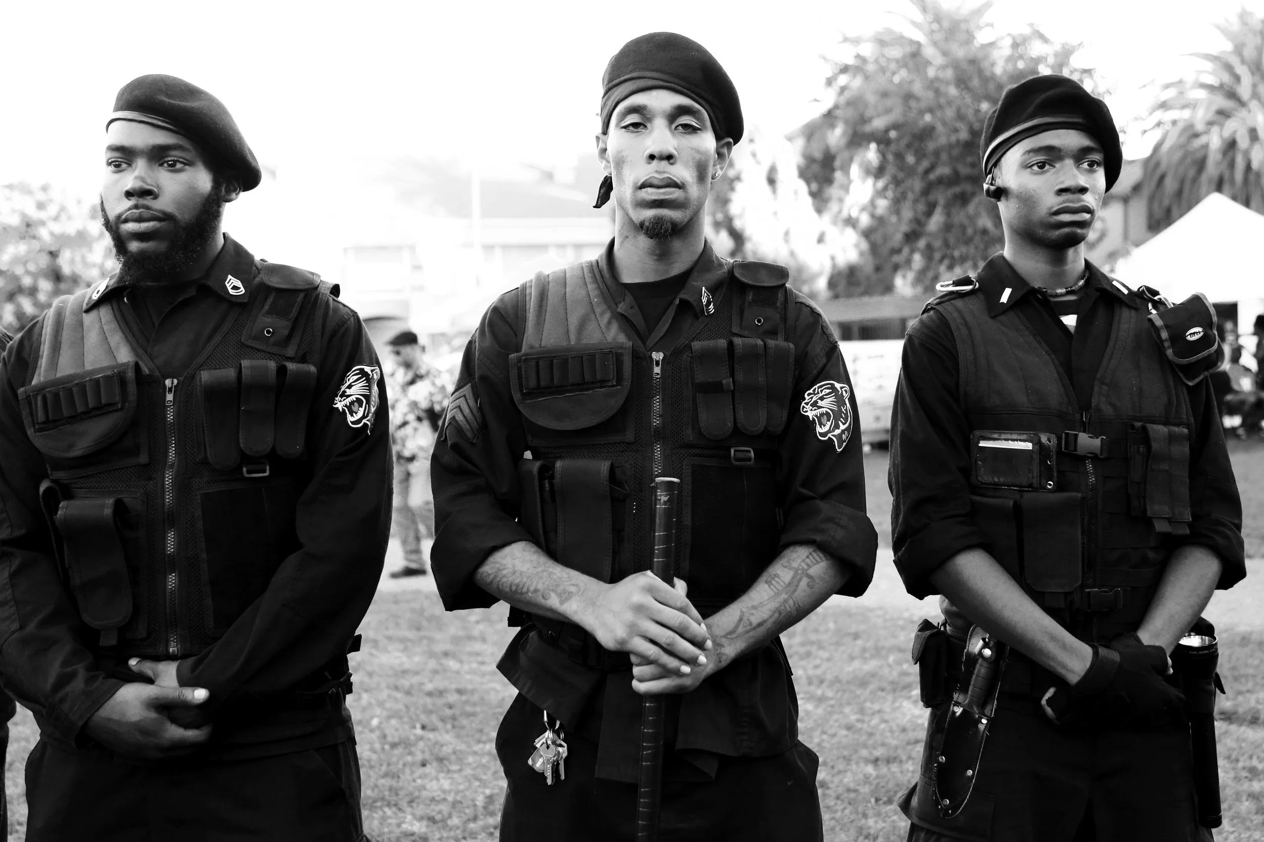 Three young men wearing black tactical gear and berets standing outdoors with trees in the background.